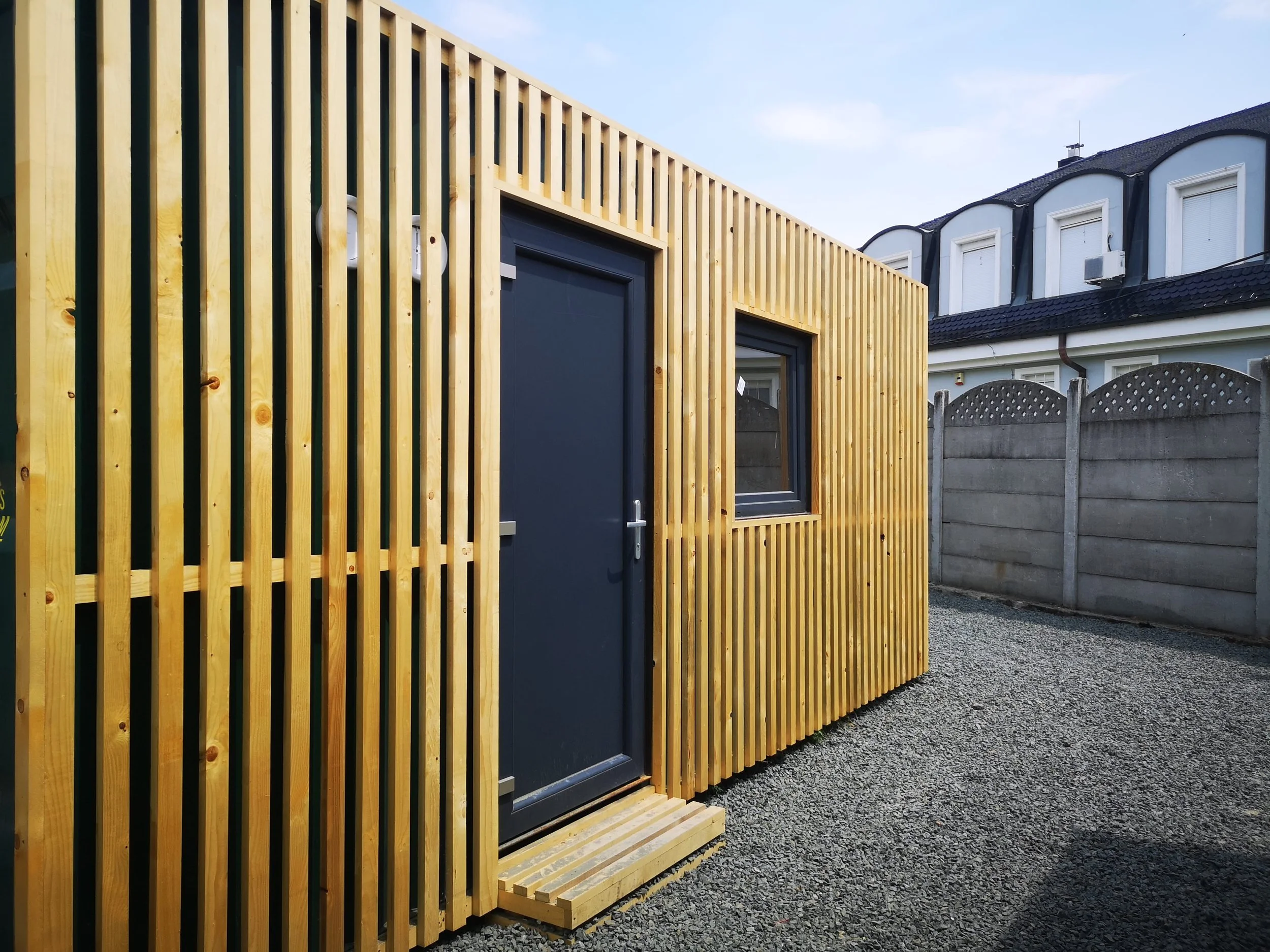 A building with a modern wooden lattice exterior, a black door, a window, and a wooden step, with a neighboring house and a concrete fence in the background.