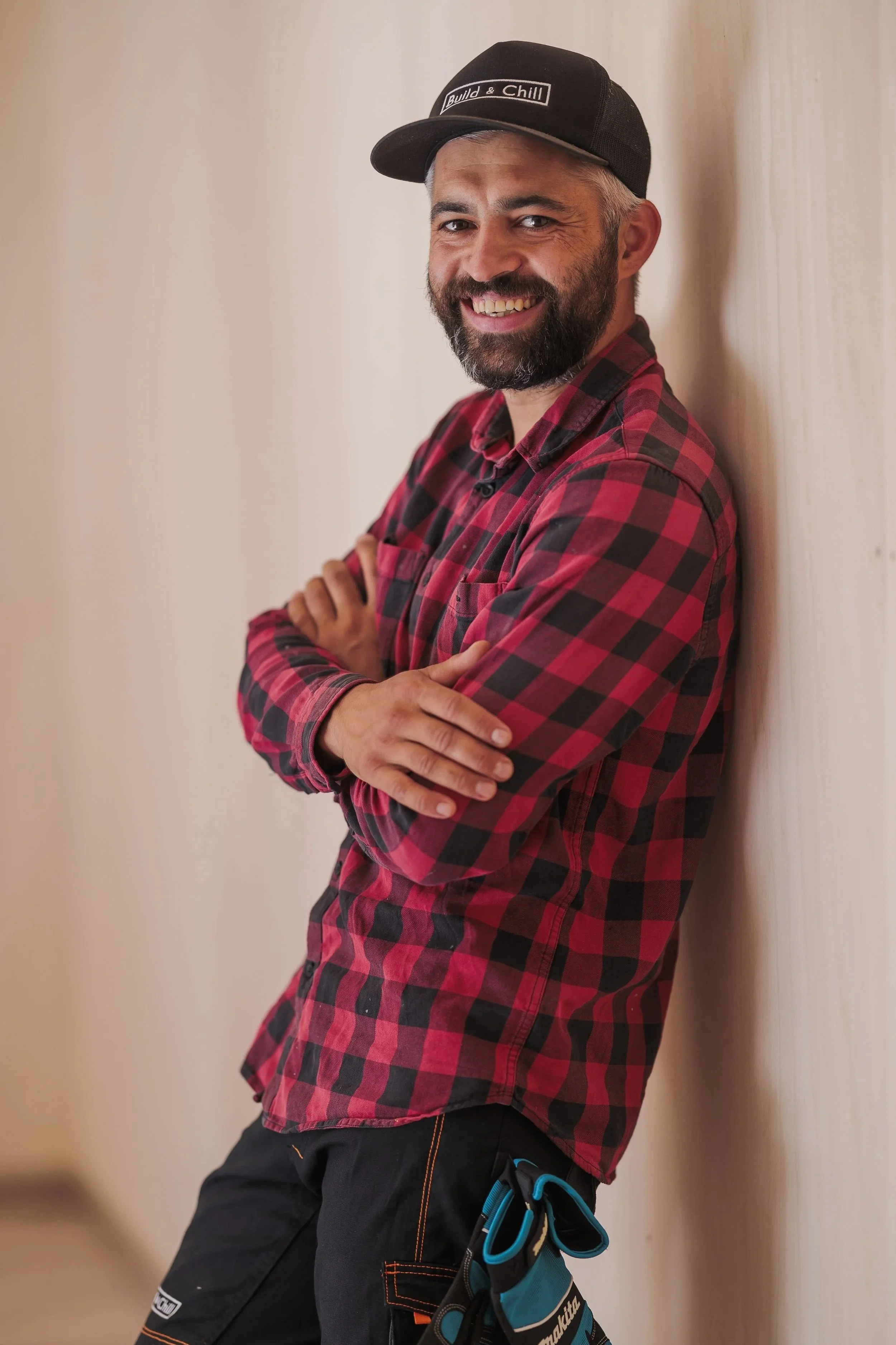 A man with gray hair and a beard leaning against a beige wall, smiling at the camera. He is wearing a black baseball cap with the words 'Build & Chill' and a red and black checkered shirt.