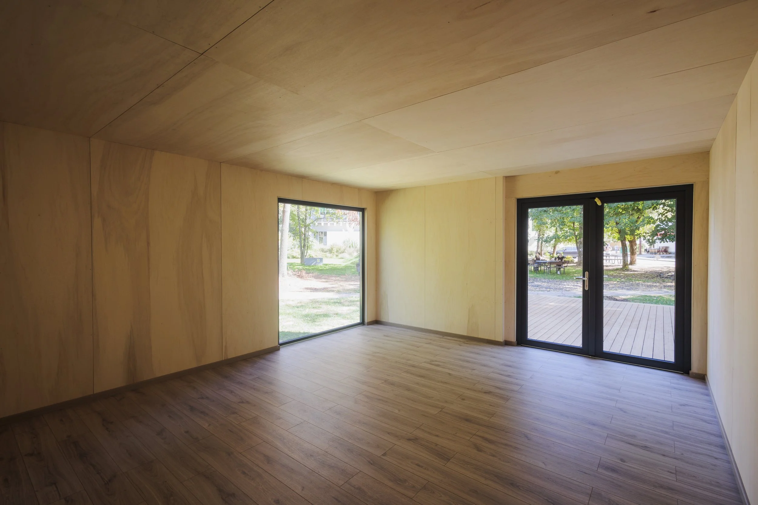 Empty room with wooden walls, ceiling, and flooring, large windows, and a glass door leading outside to a deck and a park with trees and benches.