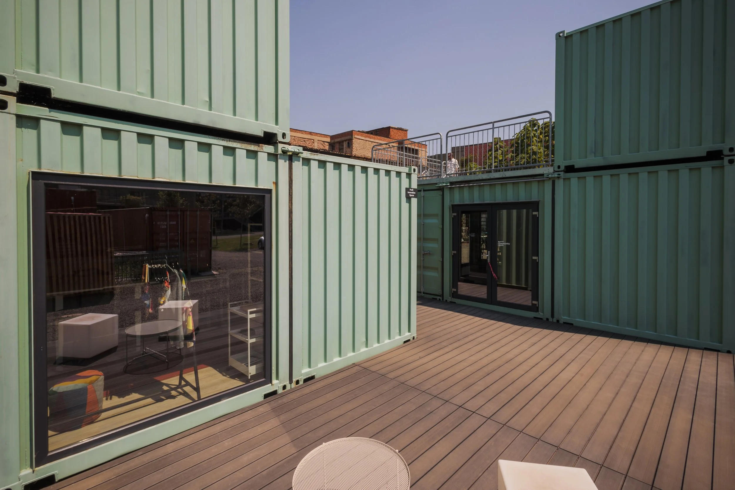 Outdoor area with green container buildings, glass windows, wooden deck flooring, and patio furniture.