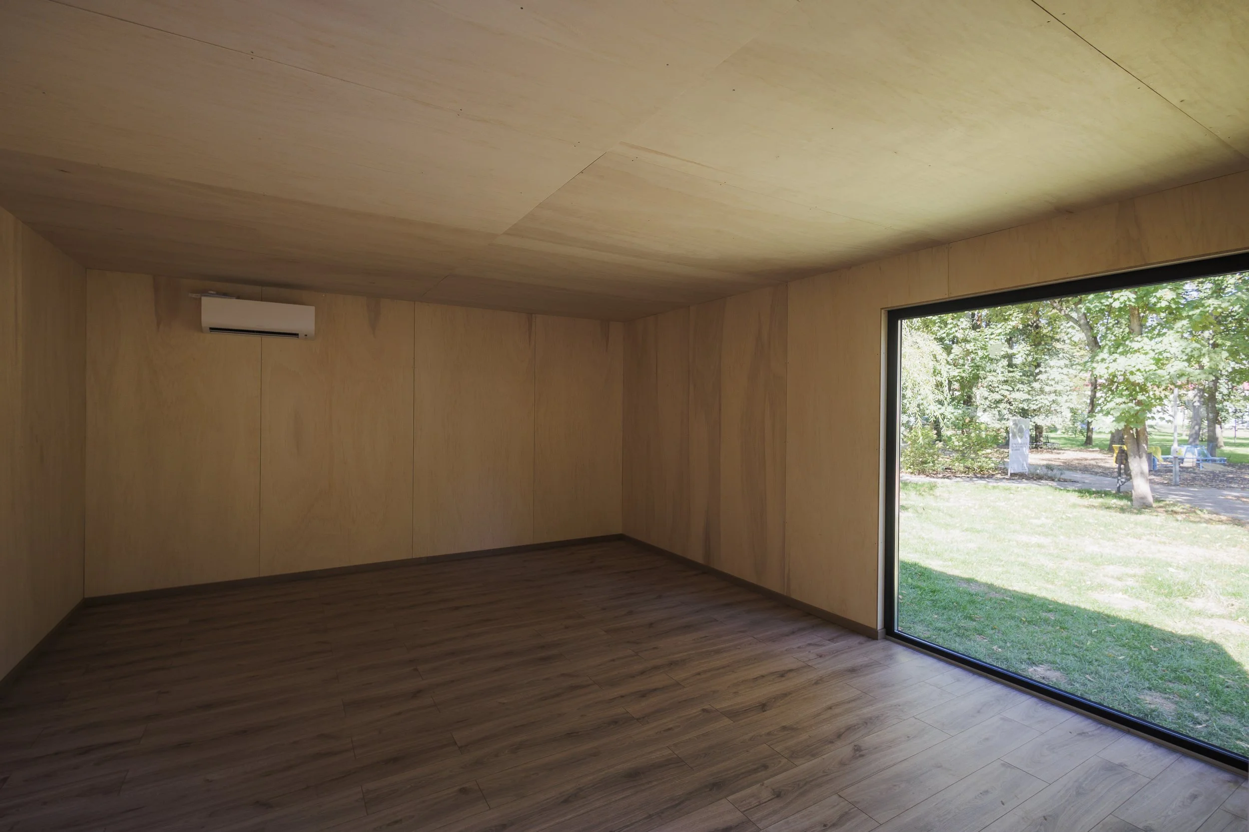 Empty room with wooden walls and ceiling, large floorboards, and a floor-to-ceiling window showing a green outdoor park with trees.