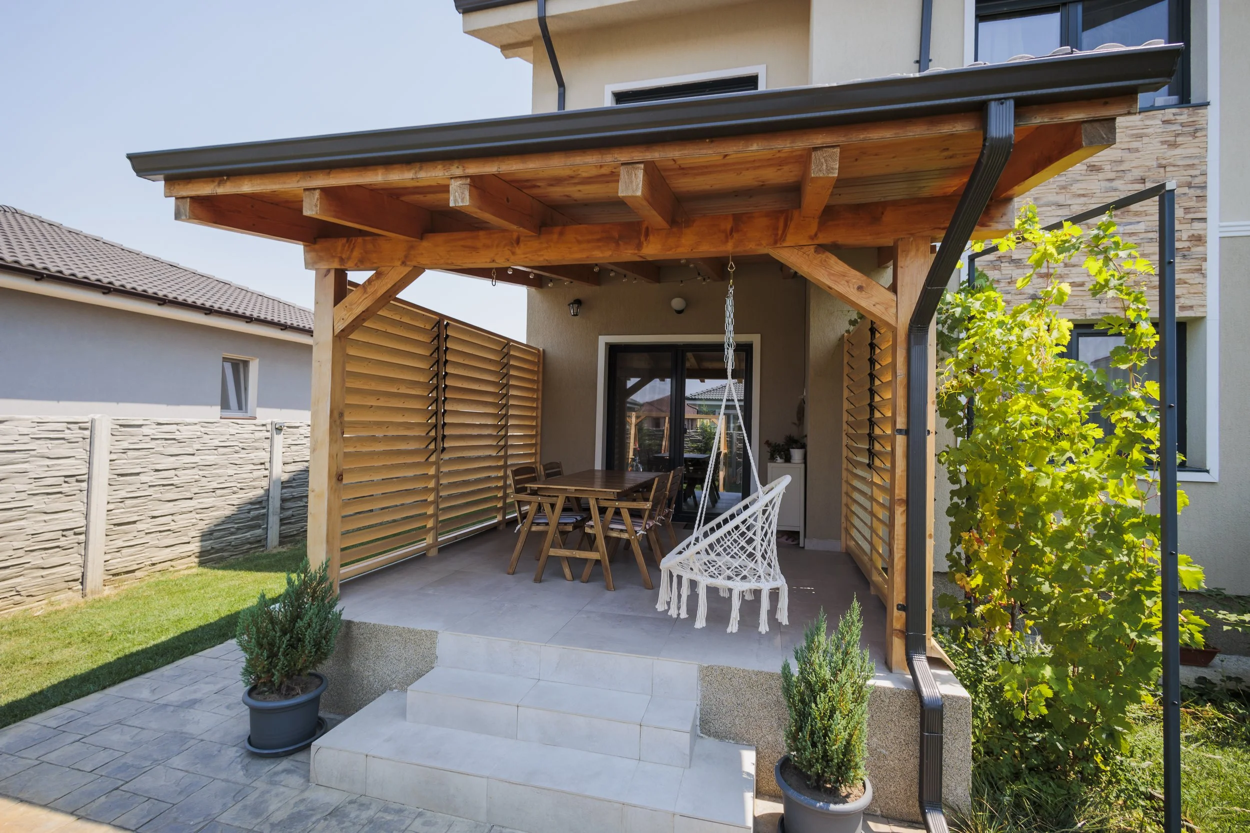 A backyard patio with a wooden pergola, outdoor dining table with chairs, a hanging swing, potted plants, and a stone wall fence, with a house in the background.