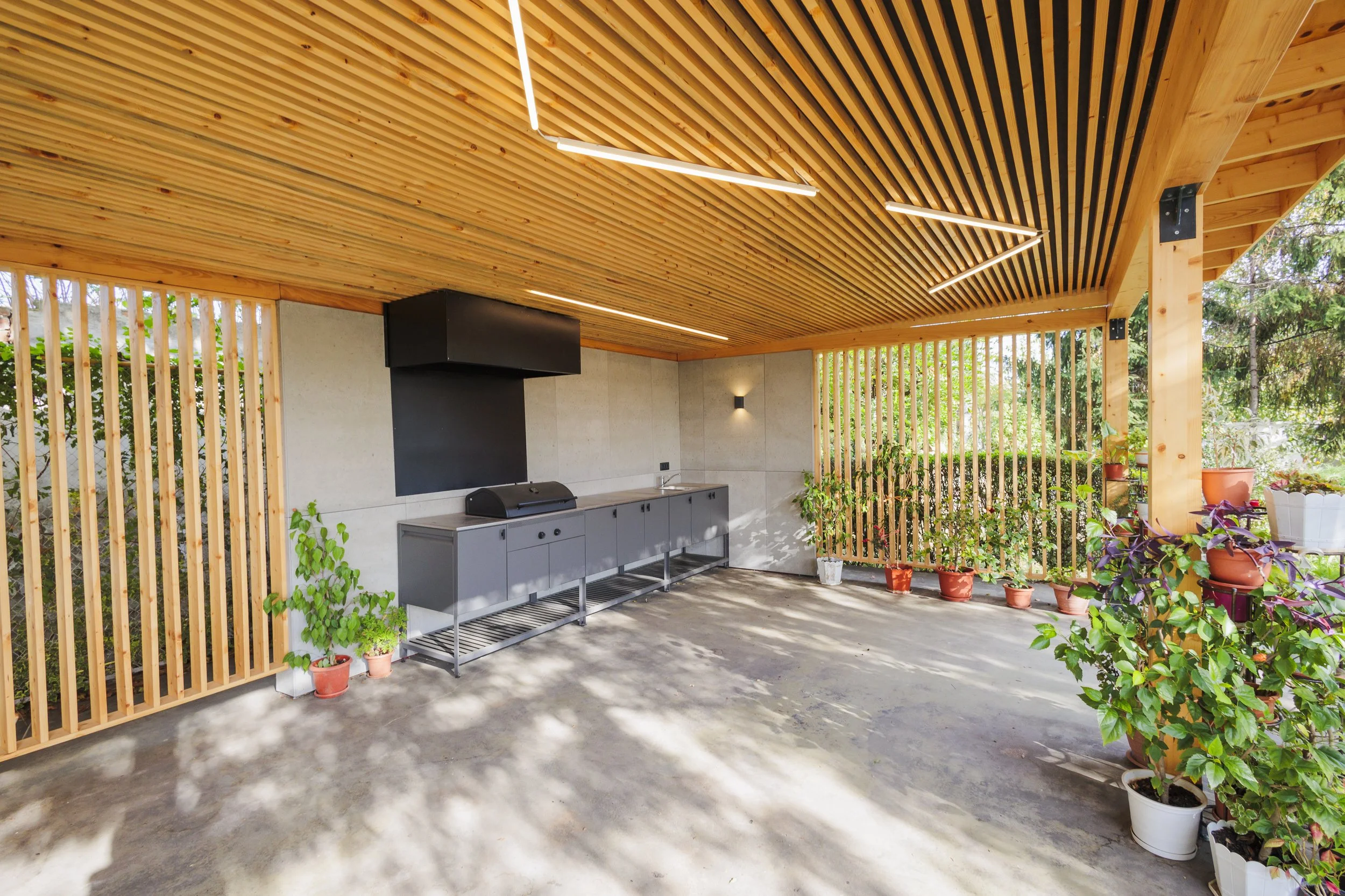 An outdoor patio area with a wood-slatted ceiling featuring modern linear LED light fixtures, a black-mounted vent or fireplace, gray cabinetry with a sink, and various potted plants along the perimeter.
