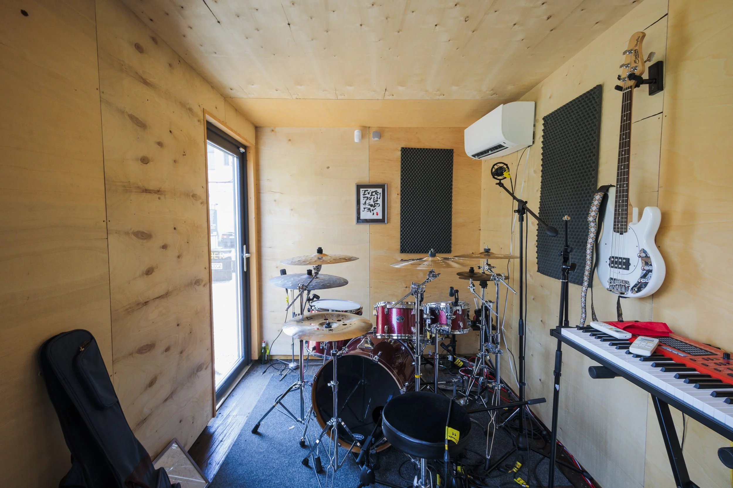 A music practice room with wooden walls and ceiling, containing a drum set, a white electric bass guitar on a stand, a keyboard, microphone, headphones, and soundproofing foam panels.