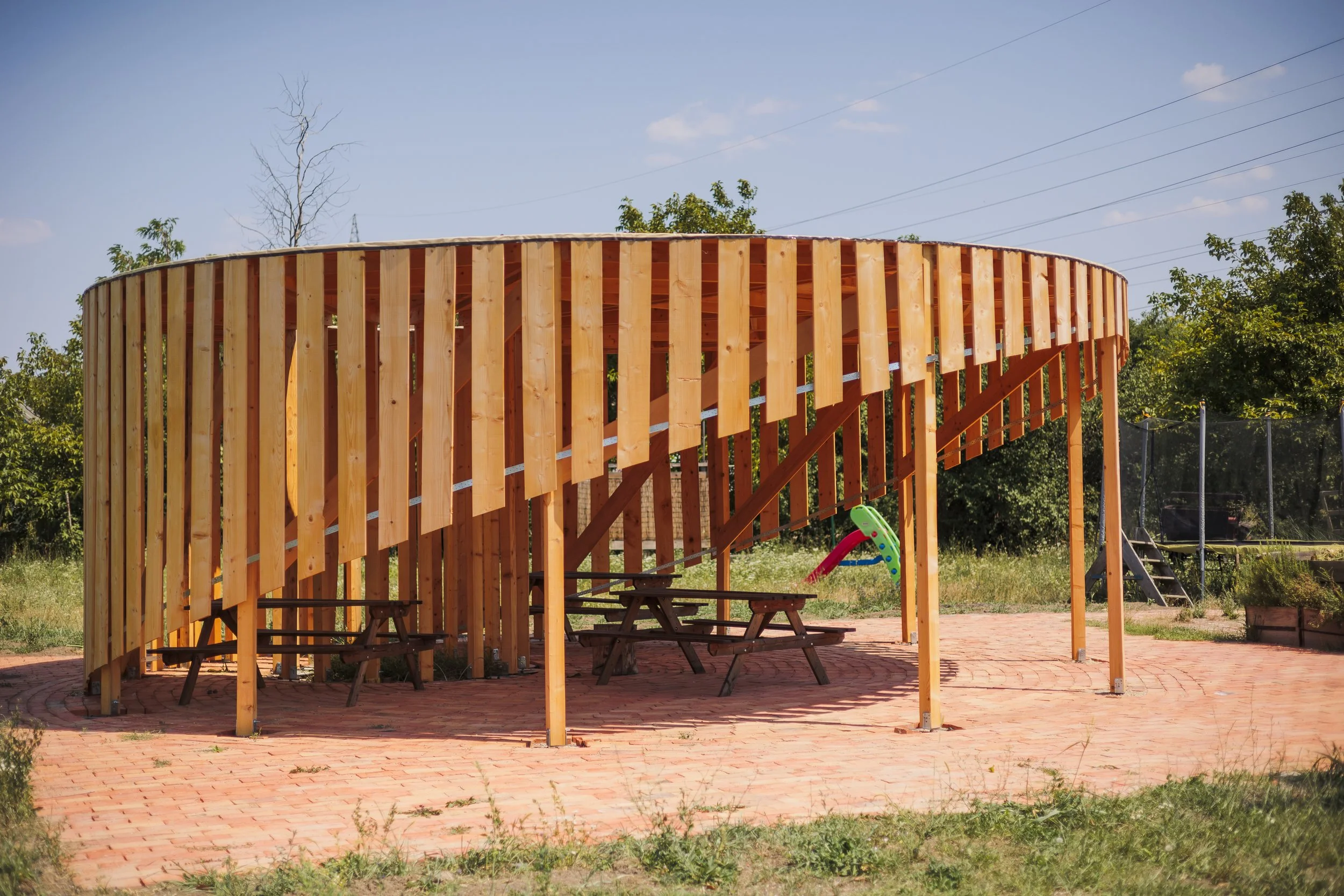 A wooden outdoor structure in a backyard, with picnic tables beneath it, a small slide, grass, trees, and a trampoline in the background, under a blue sky.