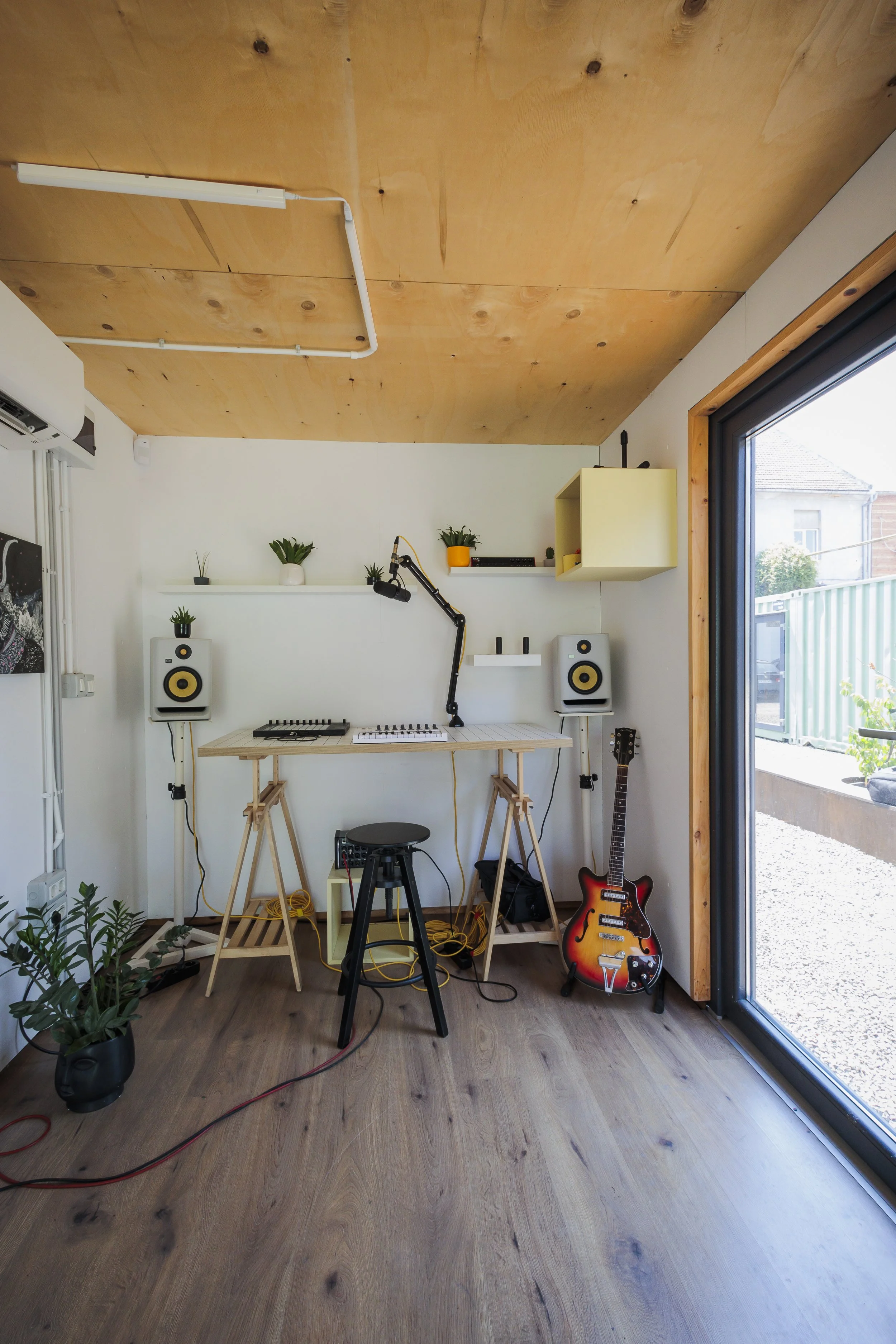 A music studio with a wooden ceiling, white walls, and a large window with a wooden frame. The room contains two white studio speakers, a guitar on the floor, a work desk with electronic music equipment, a microphone on a boom arm, potted plants on a