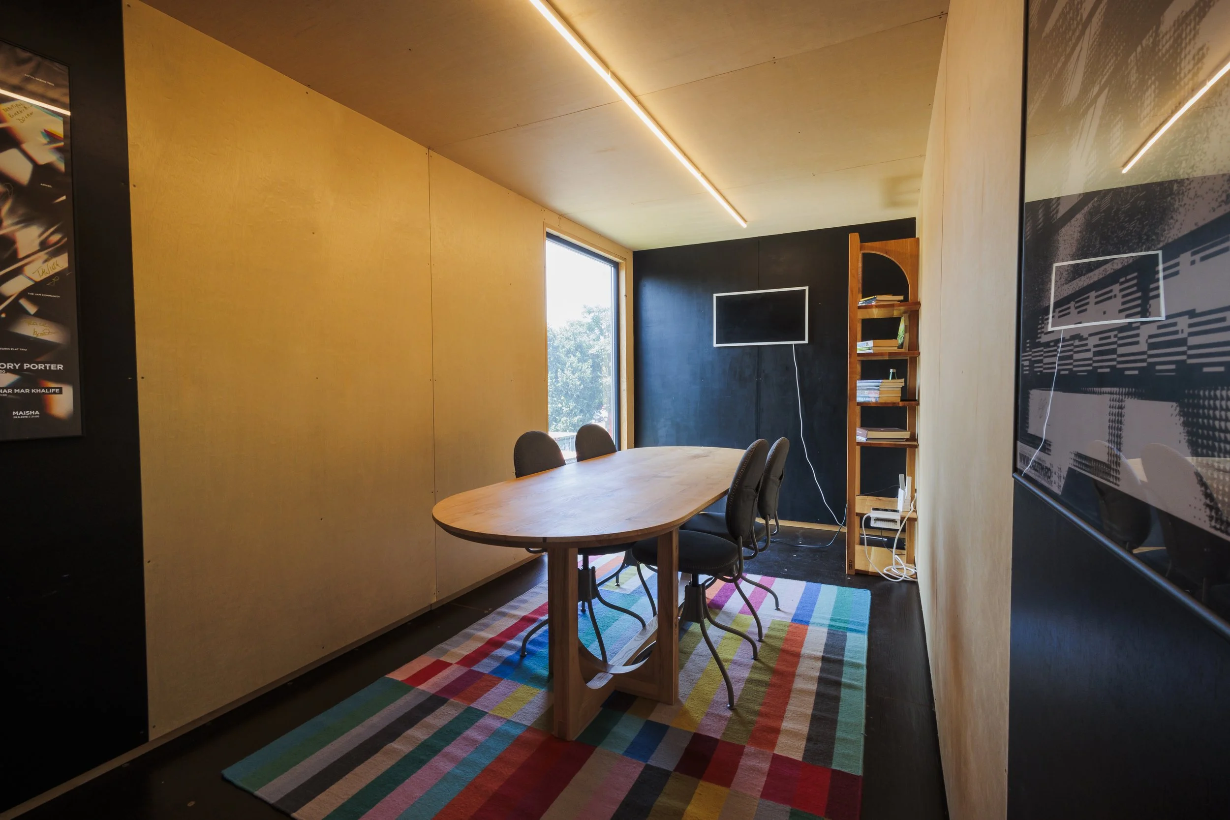 A modern conference room featuring a wooden table with black chairs, a colorful striped rug, a window with natural light, a mounted television, and a wooden bookshelf.