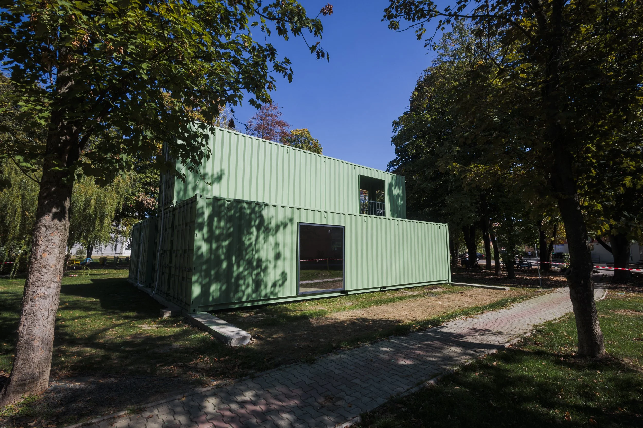 A green shipping container used as a building, located in a park with trees and paved walkways, under a blue sky.