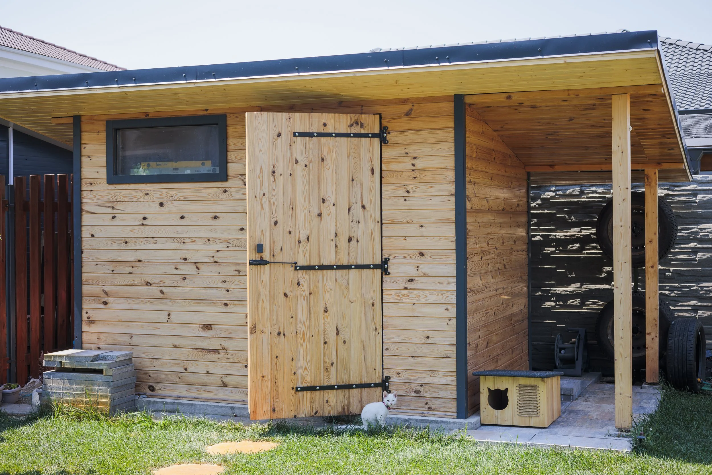 Wooden outdoor shed with a black roof, small window, and a door, situated on a grassy yard. A white cat with black markings is sitting in front of the shed near a wooden cat house.