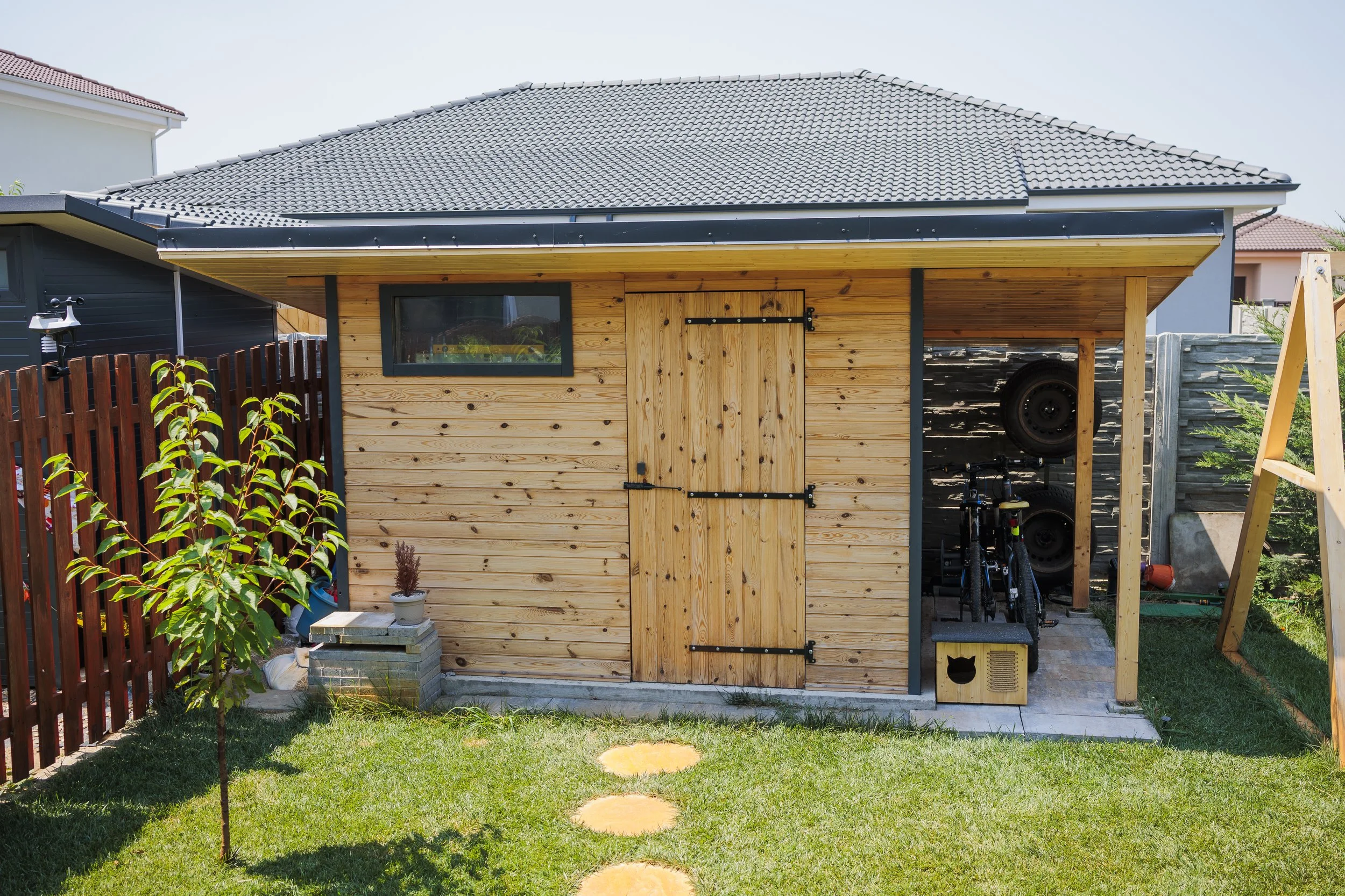 A small shed with wooden siding and a sliding barn door, situated in a backyard with a grassy lawn, a young tree, and a red wooden fence. There is a small potted plant, a stack of concrete blocks, and a black bicycle underneath the shed's covered are