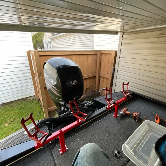 View of a boat in a garage with a Mercury outboard motor attached, red boat stands supporting it, a plastic storage bin, and a power drill on the black carpeted floor.