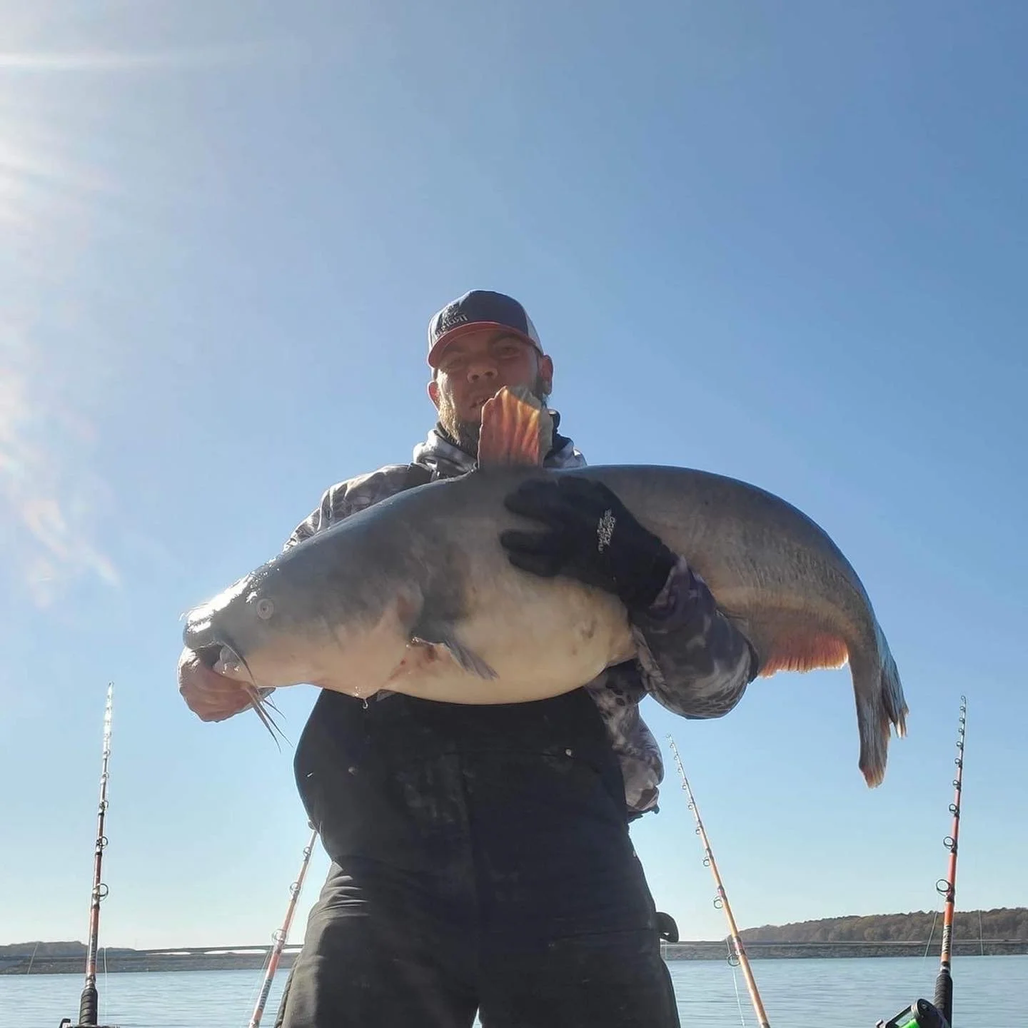 Man holding a large fish outdoors during daytime with fishing poles in the water behind him.