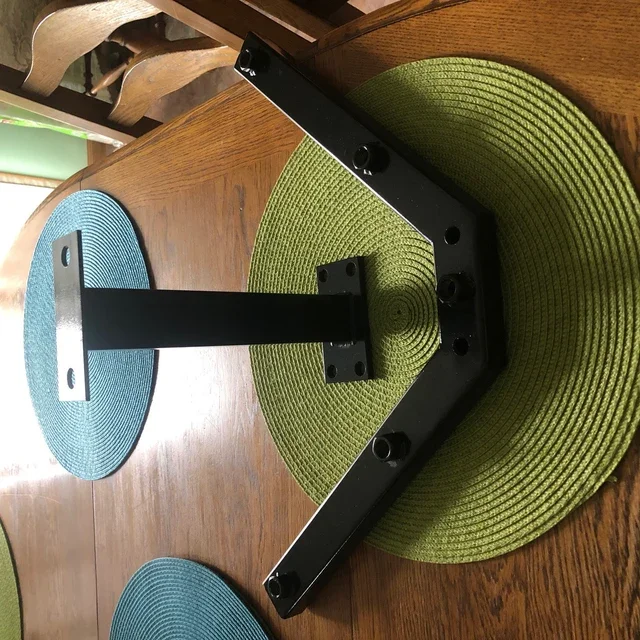 Black metal monitor stand base on a wooden table, surrounded by green and blue woven placemats.