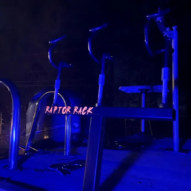 A dark gym with blue LED lighting, featuring a metal exercise machine labeled "Raptor Rack" and other fitness equipment.