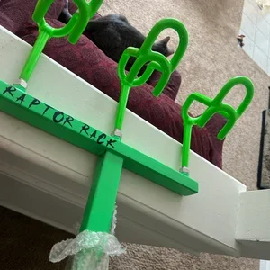 A green toy seesaw with a plastic bag hanging from one end, placed on a white surface near a purple blanket and a black tire in the background.