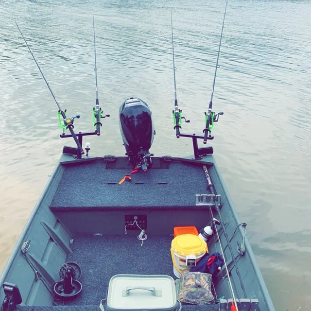 View of a fishing boat with four fishing poles mounted, an outboard motor, and various fishing gear on the deck, floating on a calm body of water.