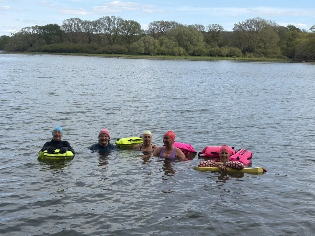 Five women are floating in a lake, each wearing swim caps and holding onto colorful flotation devices. The background features trees and a cloudy sky.
