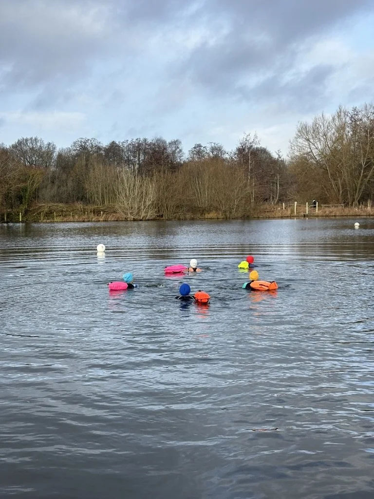Children swimming in a lake with colorful life jackets and helmets, surrounded by trees and a cloudy sky.
