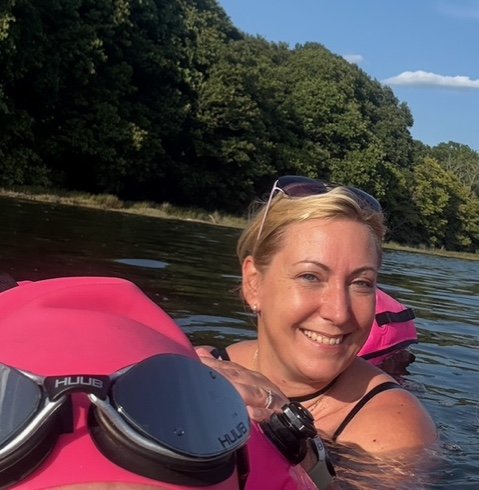 A woman smiling while floating on a river with a pink paddleboard and sunglasses on her head, surrounded by lush green trees.