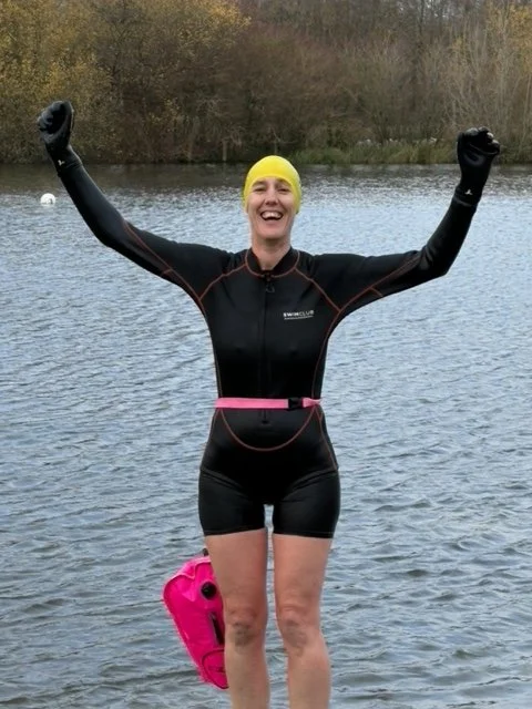 Person in a wetsuit with a yellow swim cap smiling and raising arms, standing in a lake with trees in the background.