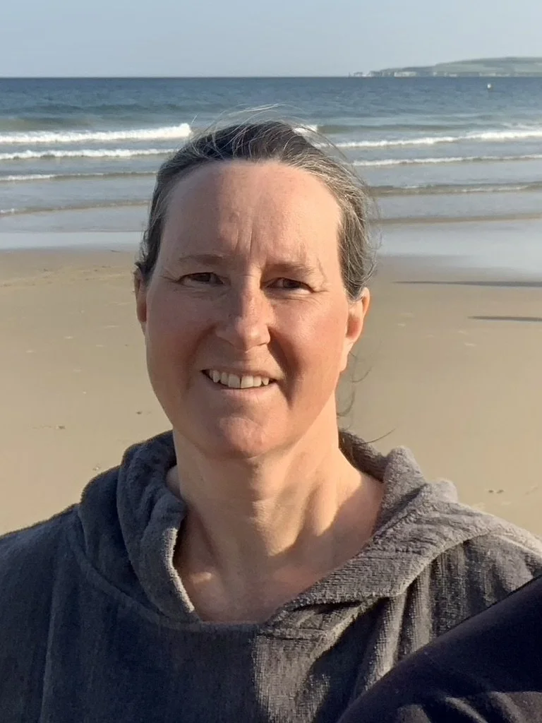 A woman taking a selfie on a beach with sand, ocean waves, and a distant land in the background.