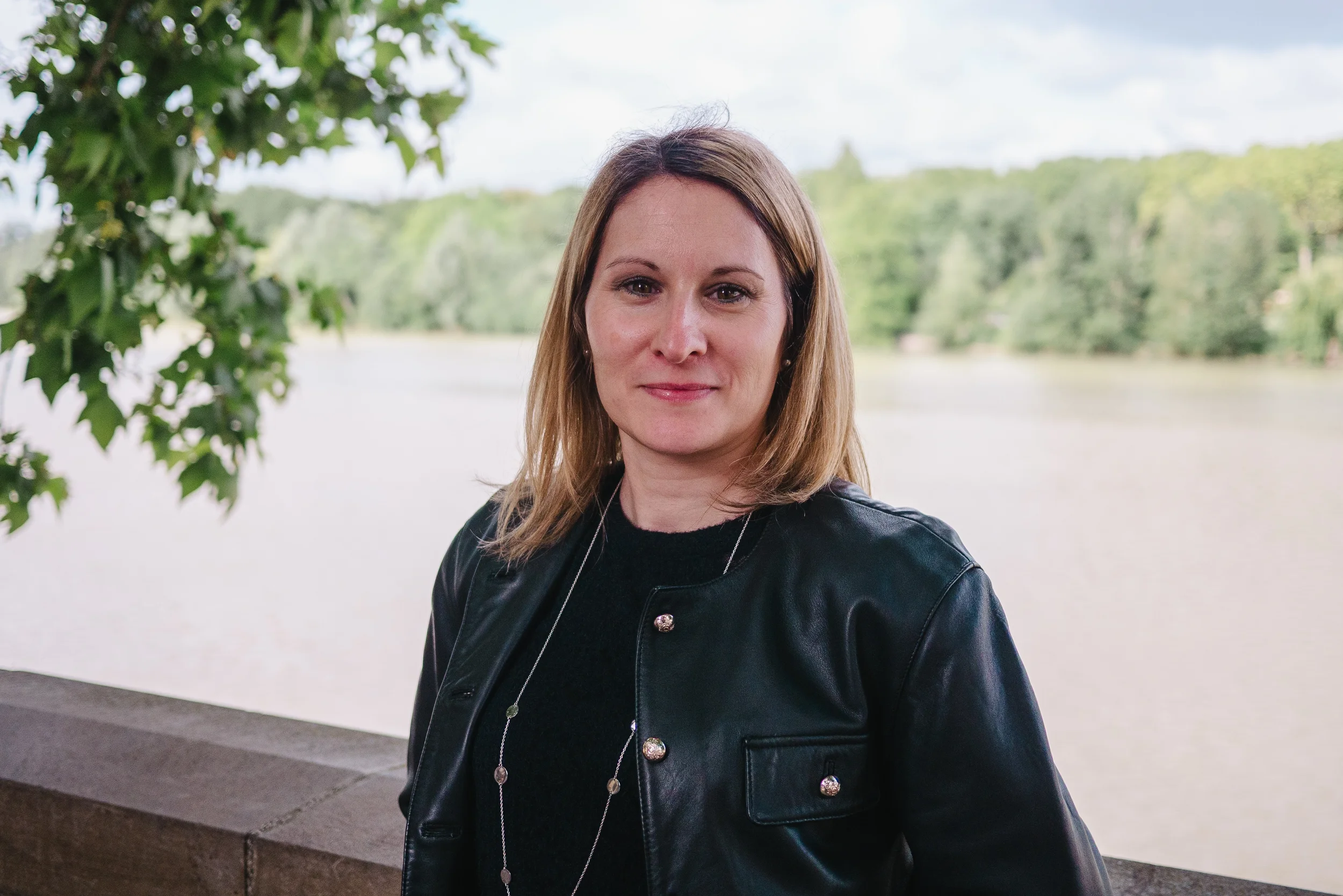 Une femme blonde portant une veste en cuir noire se tient près d'un lac avec un fond de forêt verdoyante, sous un ciel partiellement nuageux.