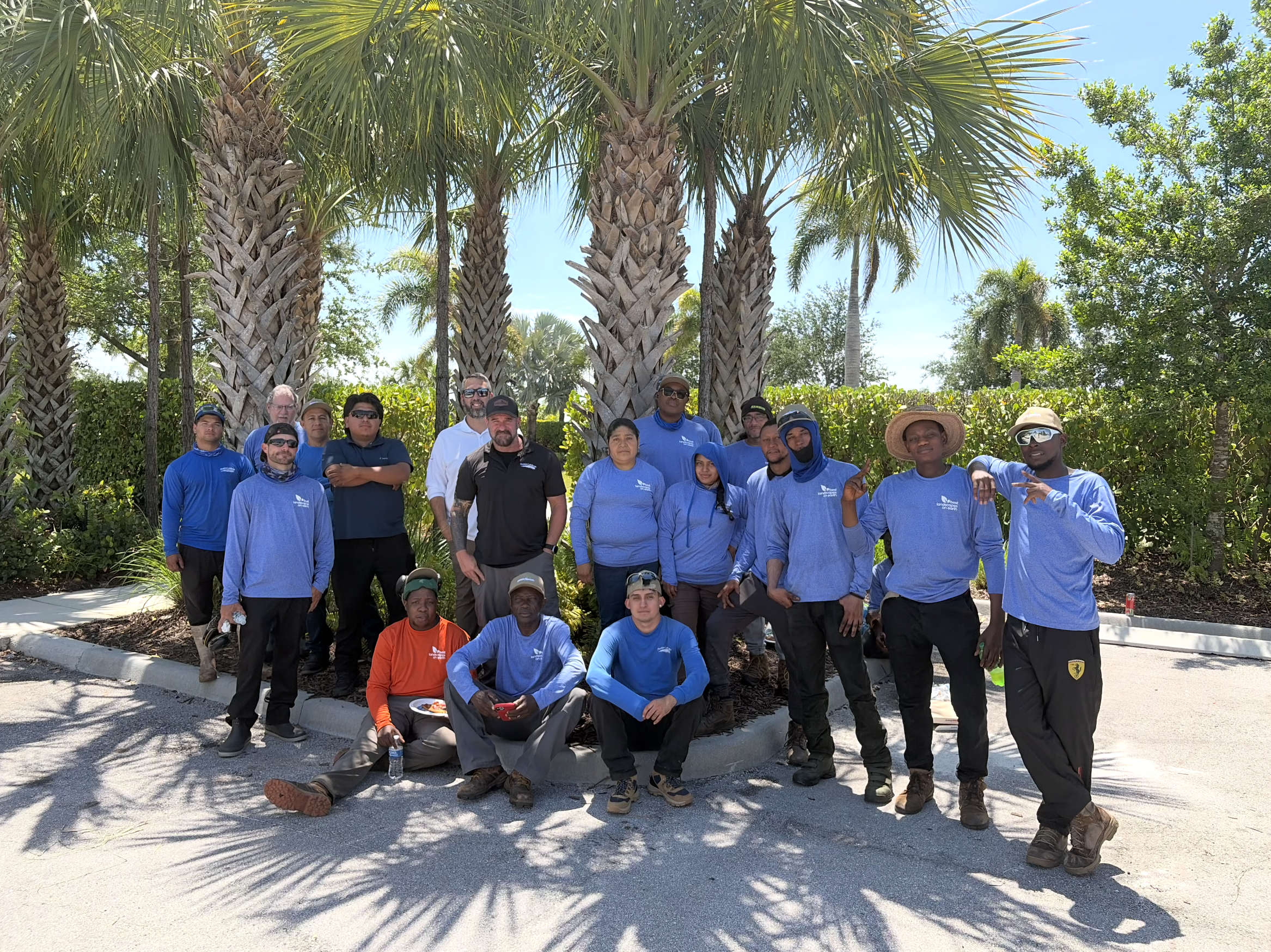 A group of diverse people posing outdoors in front of palm trees under a bright blue sky. They are wearing casual clothing, with some in matching blue shirts, and include men and women of various ages and backgrounds.