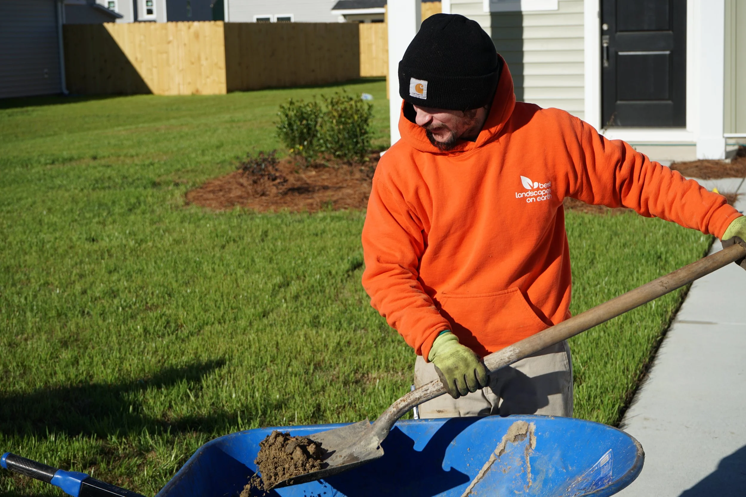 A man wearing an orange hoodie with the logo 'Best Landscapes on Earth' and a black beanie is working on landscaping by shoveling dirt into a blue wheelbarrow in a backyard with green grass, a small bush, and a wooden fence.