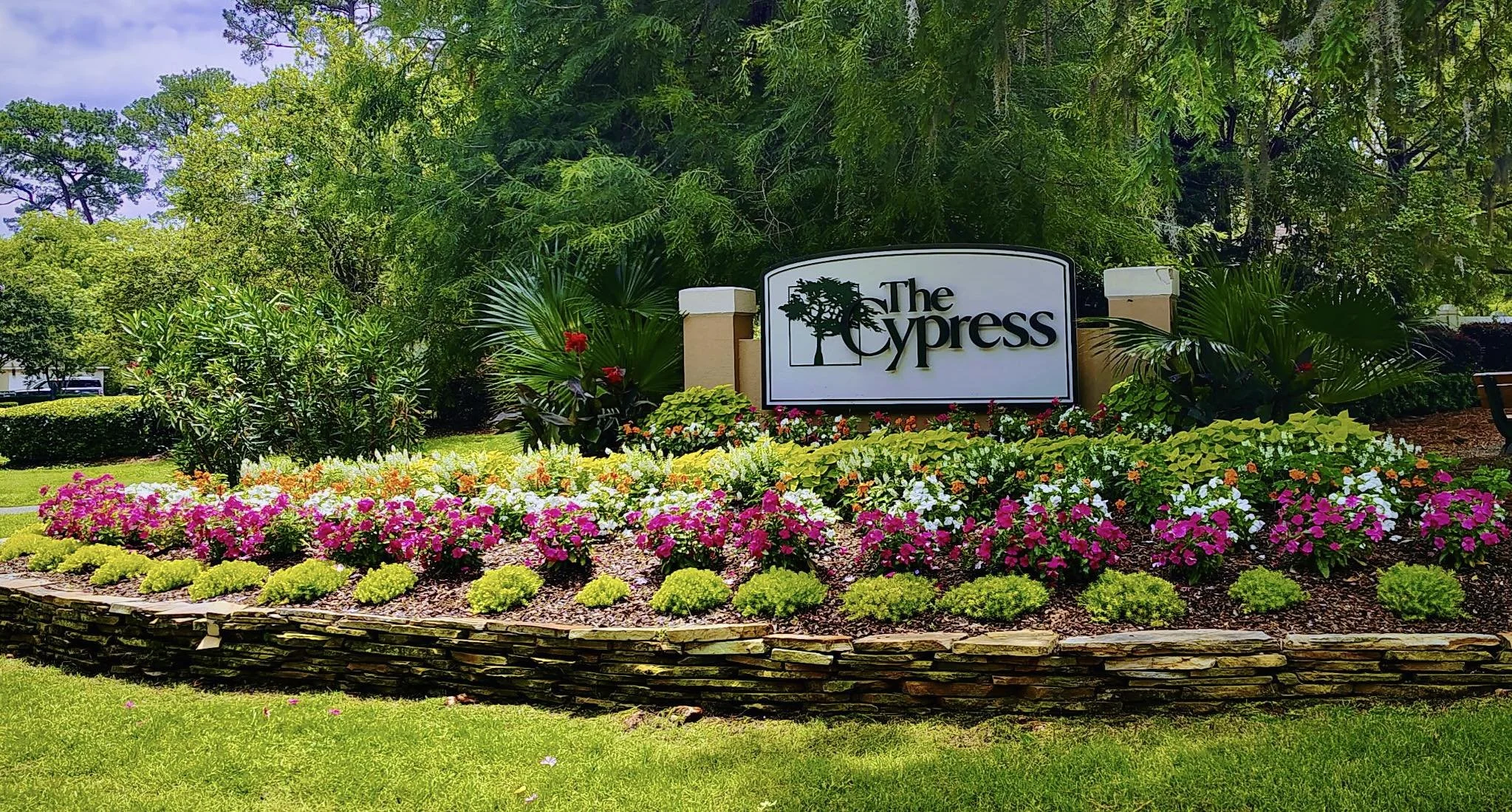A landscaped garden with a variety of colorful flowers and green bushes surrounding a sign that reads 'The Cypress' with a tree icon. Tall trees and clear sky in the background.