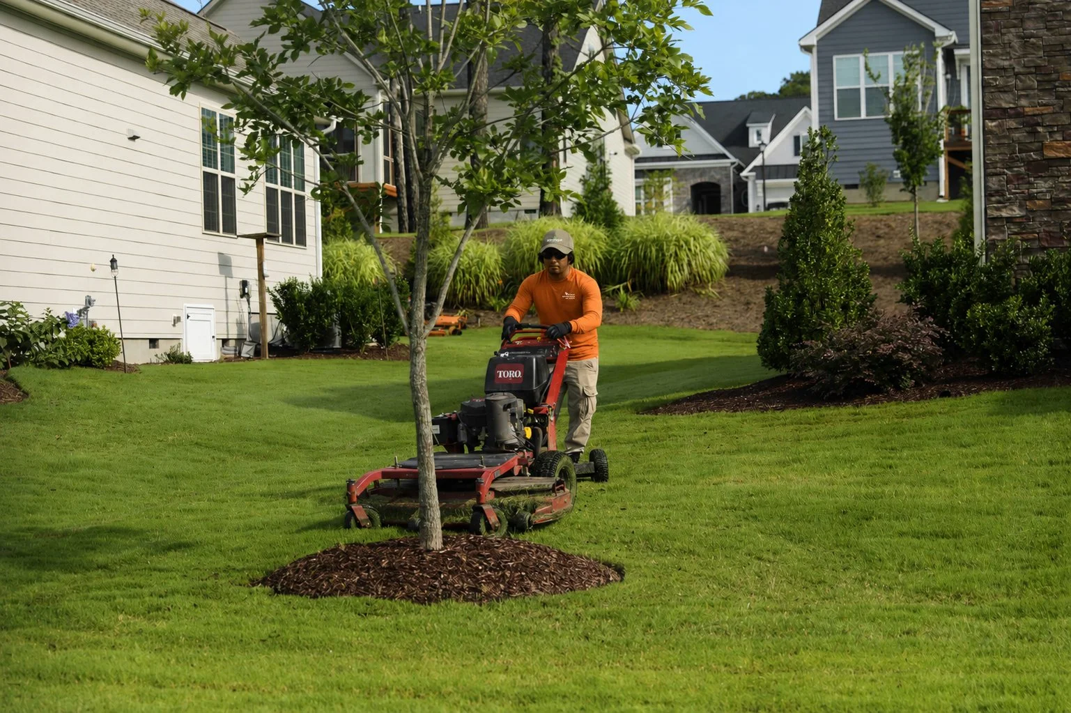 A man in an orange long-sleeve shirt and tan pants mowing a well-maintained grassy yard with a Toro riding mower around a tree with mulch at its base. Houses with landscaping and trees are in the background.