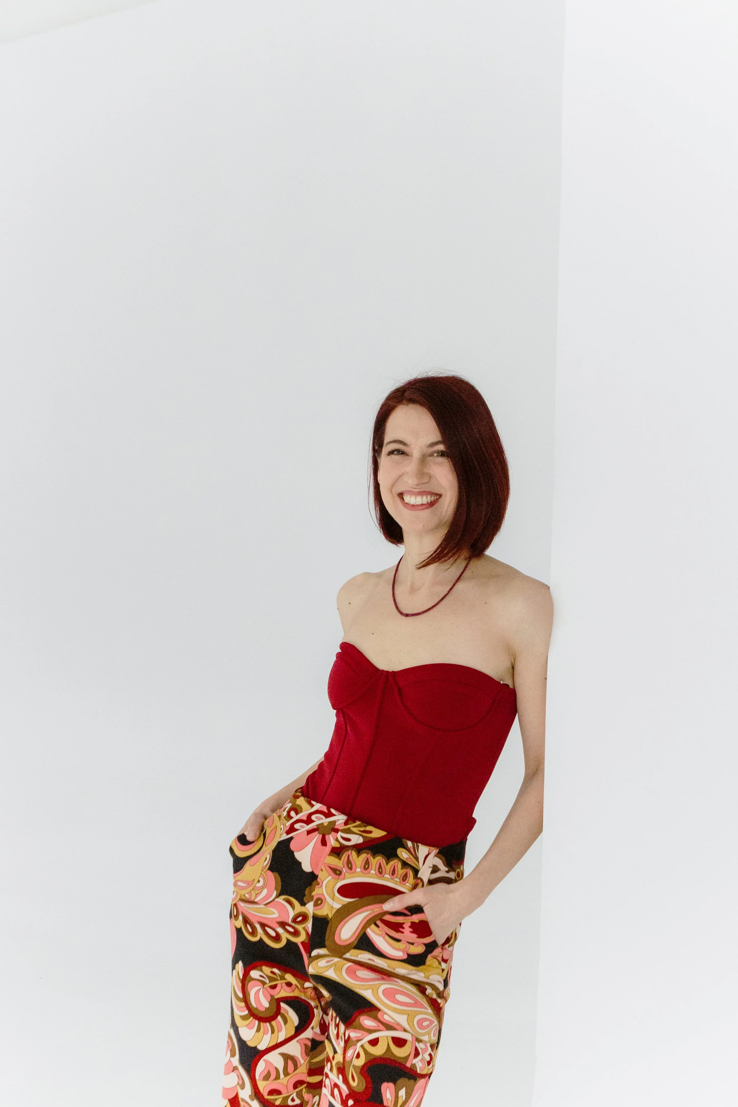 A woman with shoulder-length red hair, smiling, wearing a red strapless top, a necklace, and colorful patterned pants, standing against a white wall.