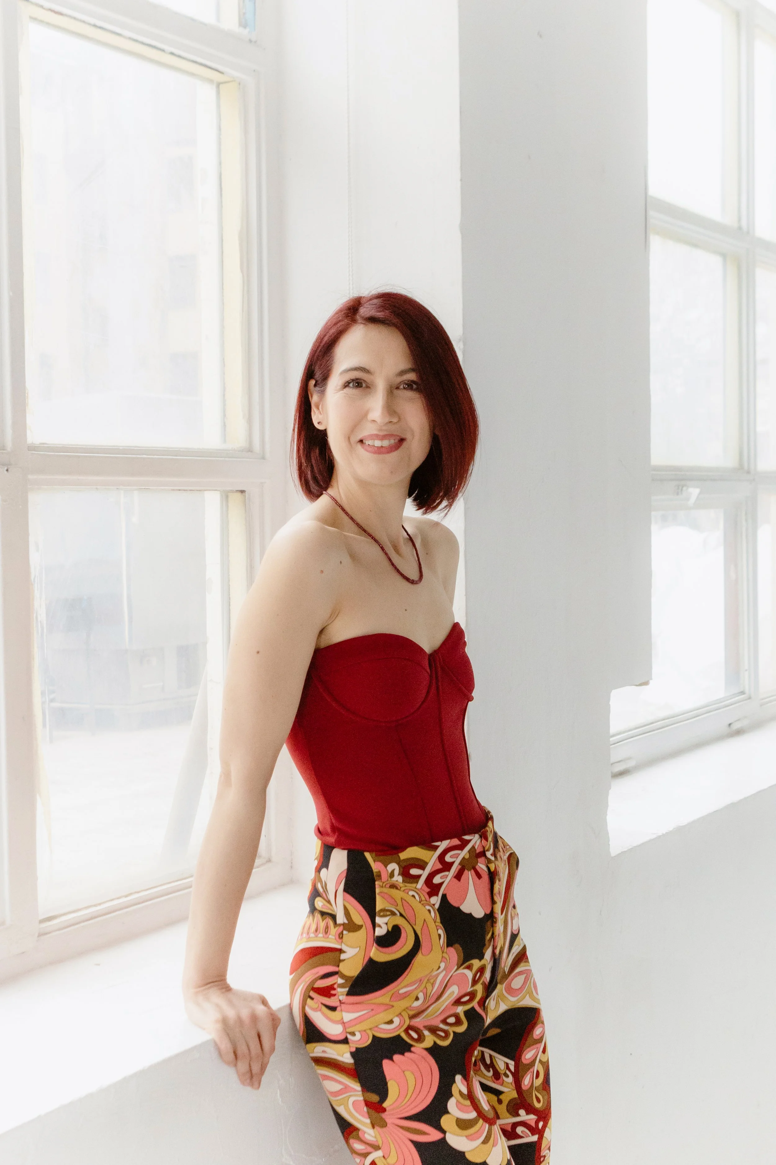 A woman with red hair smiles at the camera, wearing a strapless red top and patterned pants, standing near a white wall with large windows in the background.