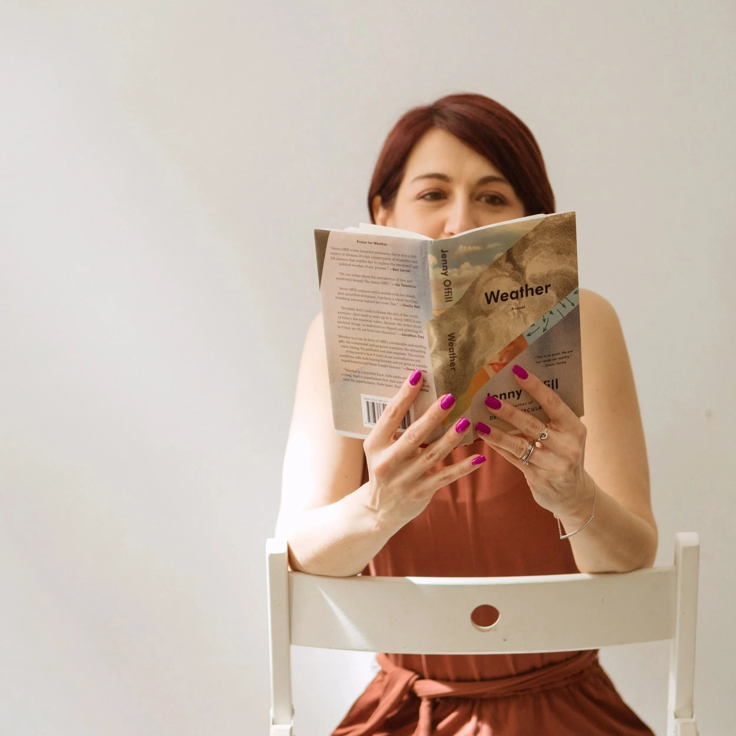 A woman with read hair is sitting on a chair and reading Jenny Offil's Weather. Her face is not fully visible behind the book.