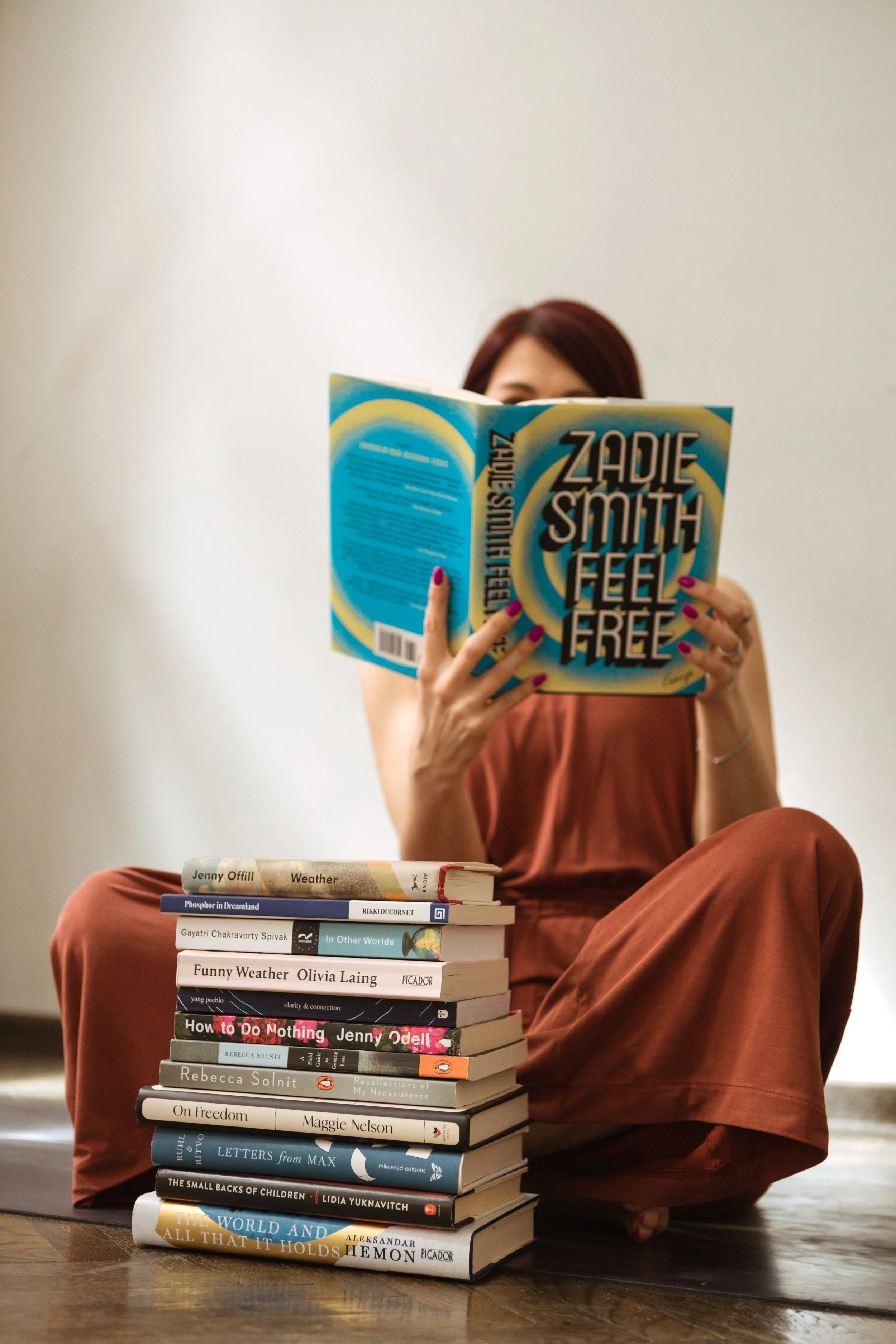 A woman with reddish-brown hair reading a book titled 'Zadie Smith Feel Free' while sitting cross-legged on the floor. There is a stack of various books on the floor in front of her, and she is wearing a brown sleeveless outfit and painted fingernails.