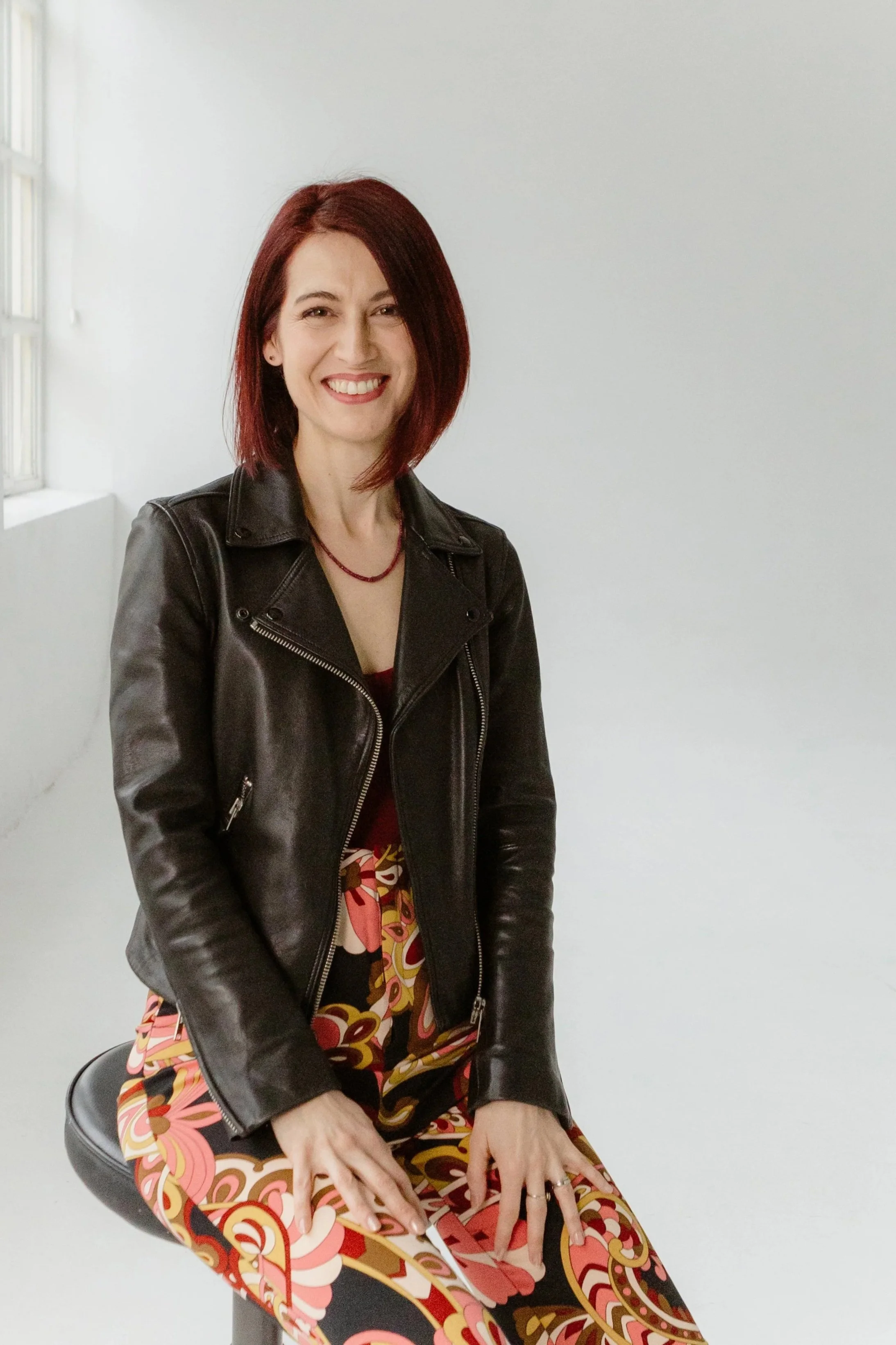 A woman with shoulder-length red hair smiling, wearing a black leather jacket, a red beaded necklace, and a colorful patterned skirt, sitting on a black stool in a minimalist room with white walls and a window.