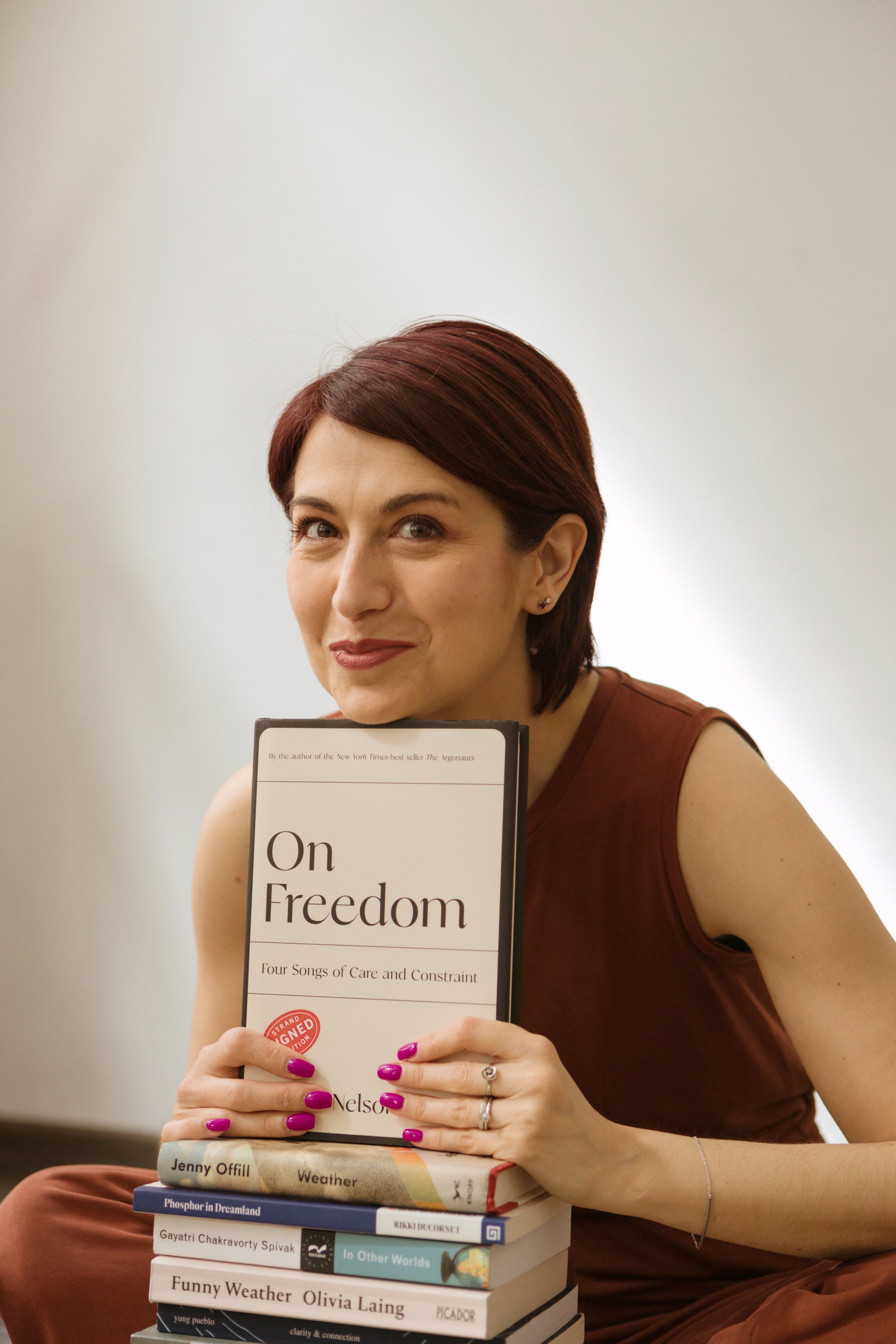 A woman with short, reddish-brown hair and purple nail polish sitting on the floor holding a hardcover book titled "On Freedom." She is wearing a brown sleeveless top and has a silver ring on her right hand. Several other books are stacked beside her.