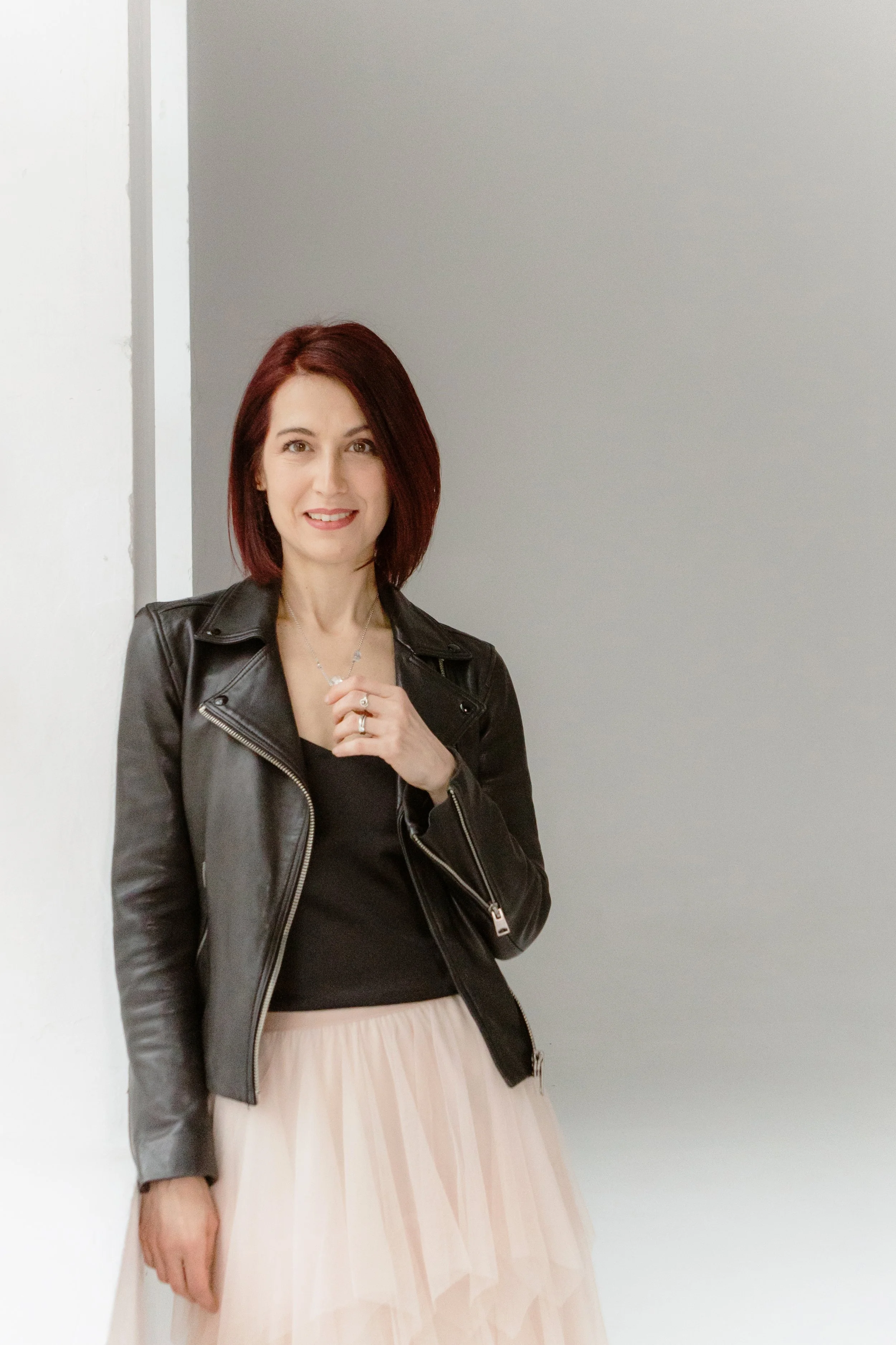 A woman with shoulder-length red hair wearing a black leather jacket, black top, and a long beige skirt, standing against a plain wall.