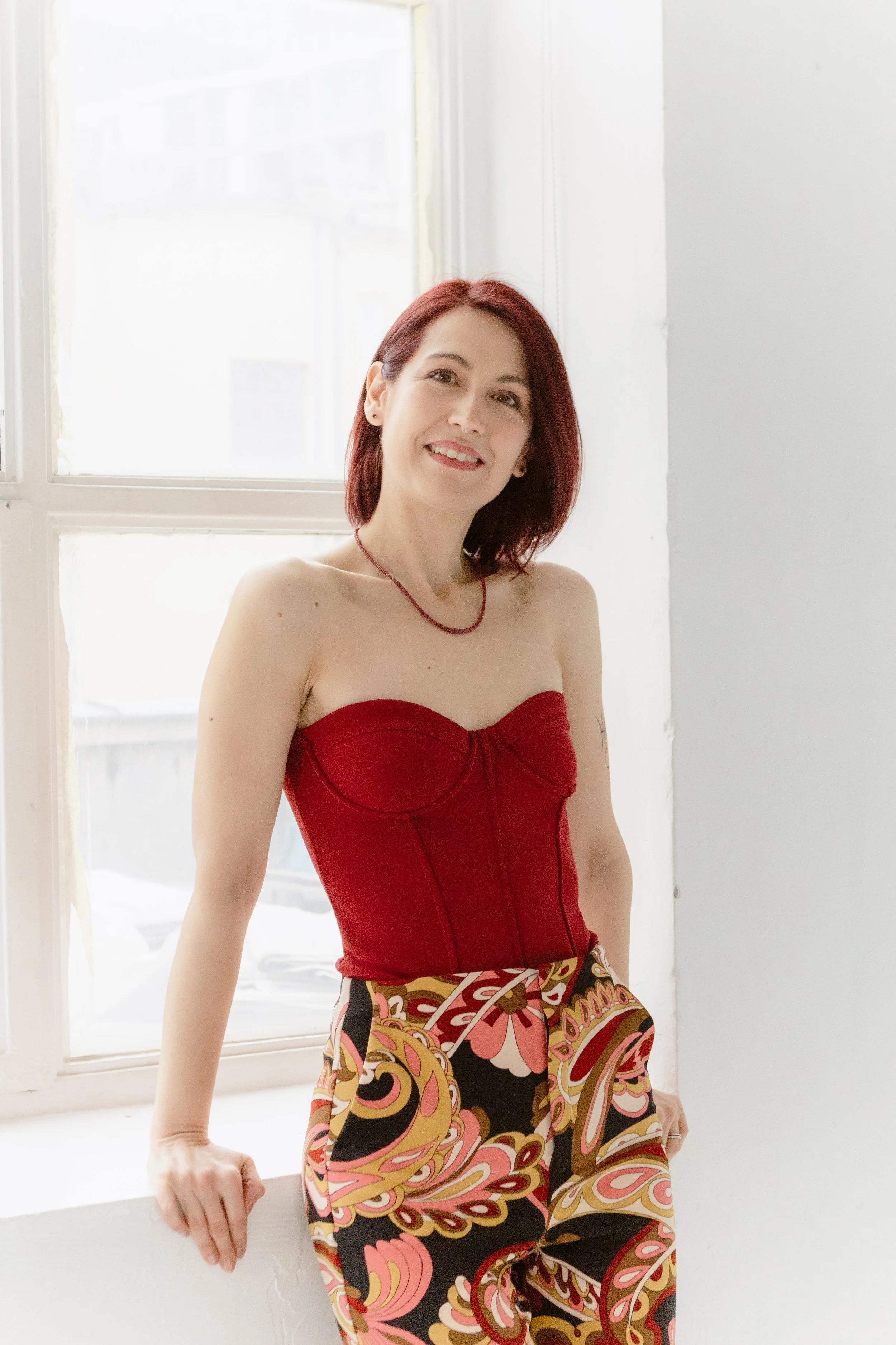 A woman with shoulder-length red hair smiling in front of a white window, wearing a strapless red top, colorful patterned pants, and a matching necklace, sitting on a white ledge.