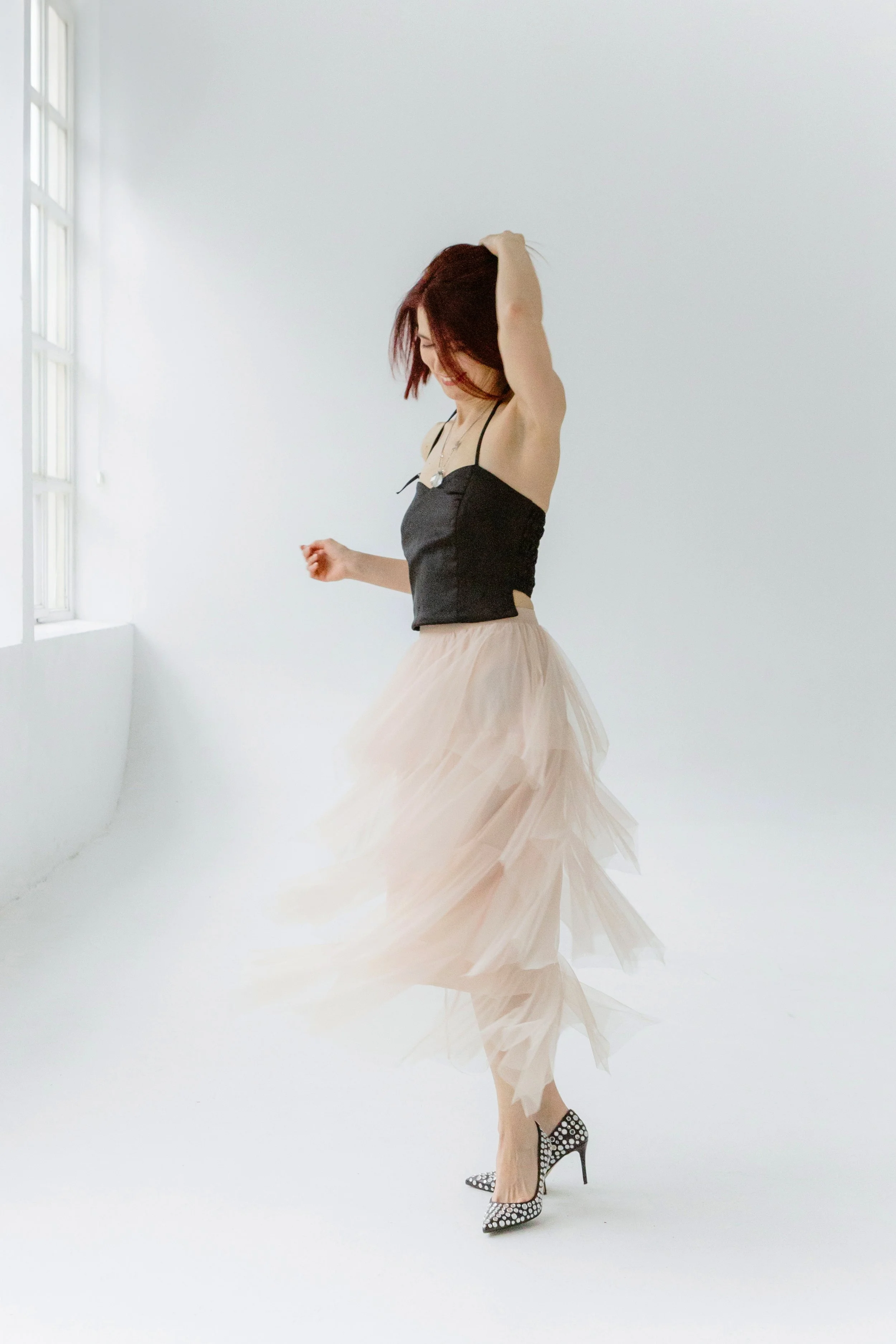 A woman with short red hair dancing in a bright, minimalistic room. She is wearing a black strap top, a flowing beige tulle skirt, and black polka dot high heels.