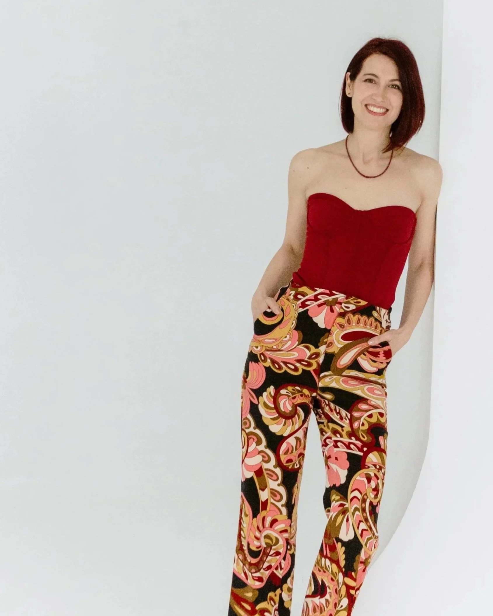 A woman with red hair wearing a red strapless top and colorful patterned pants, smiling and leaning against a white wall.
