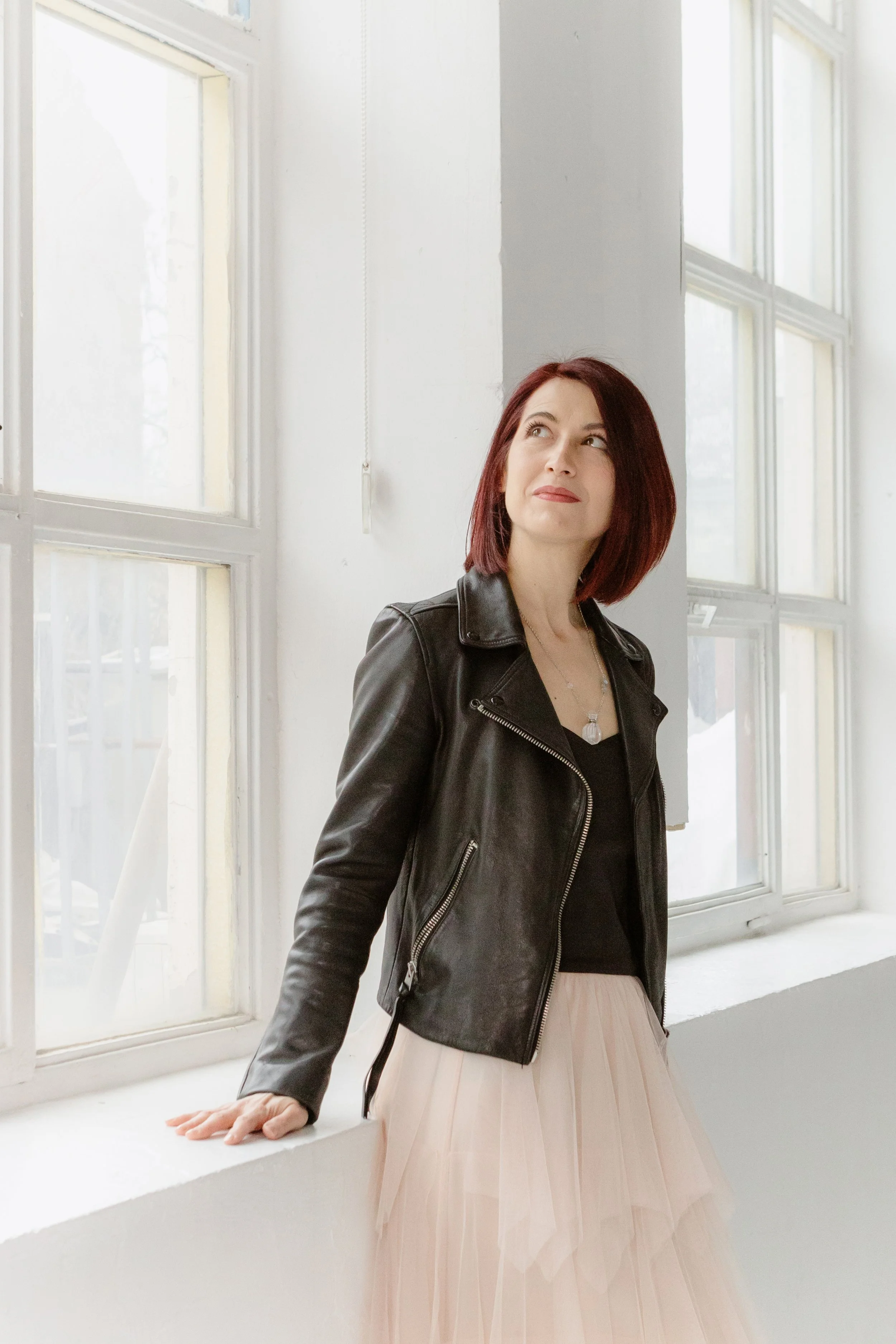 A woman with red hair wearing a black leather jacket and a light pink tulle skirt, standing near a white windowsill in a bright room, looking up and to the side.