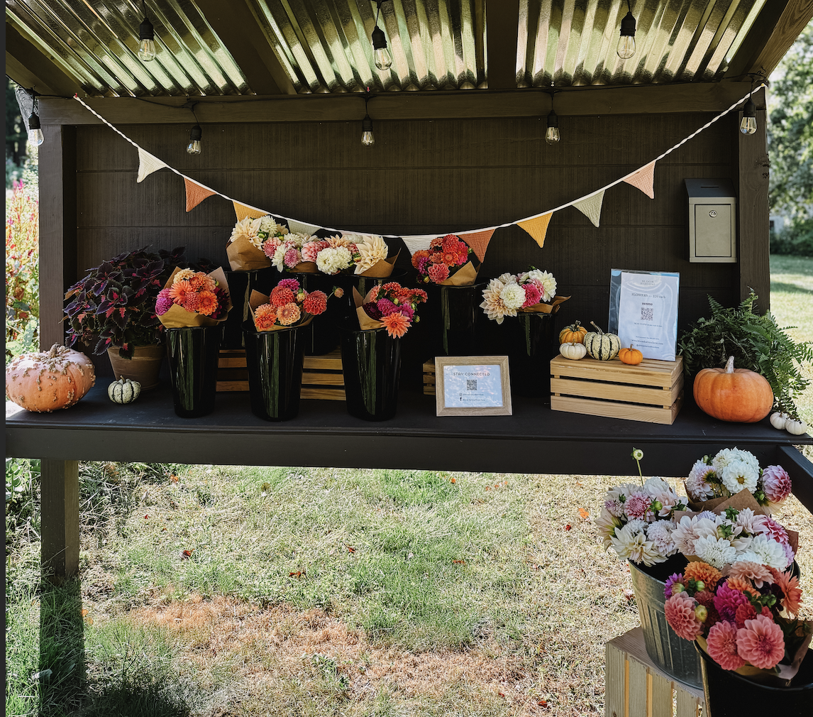 Outdoor display with colorful flower bouquets, pumpkins, and autumn decorations under a metal roof with string lights and a burlap bunting banner.