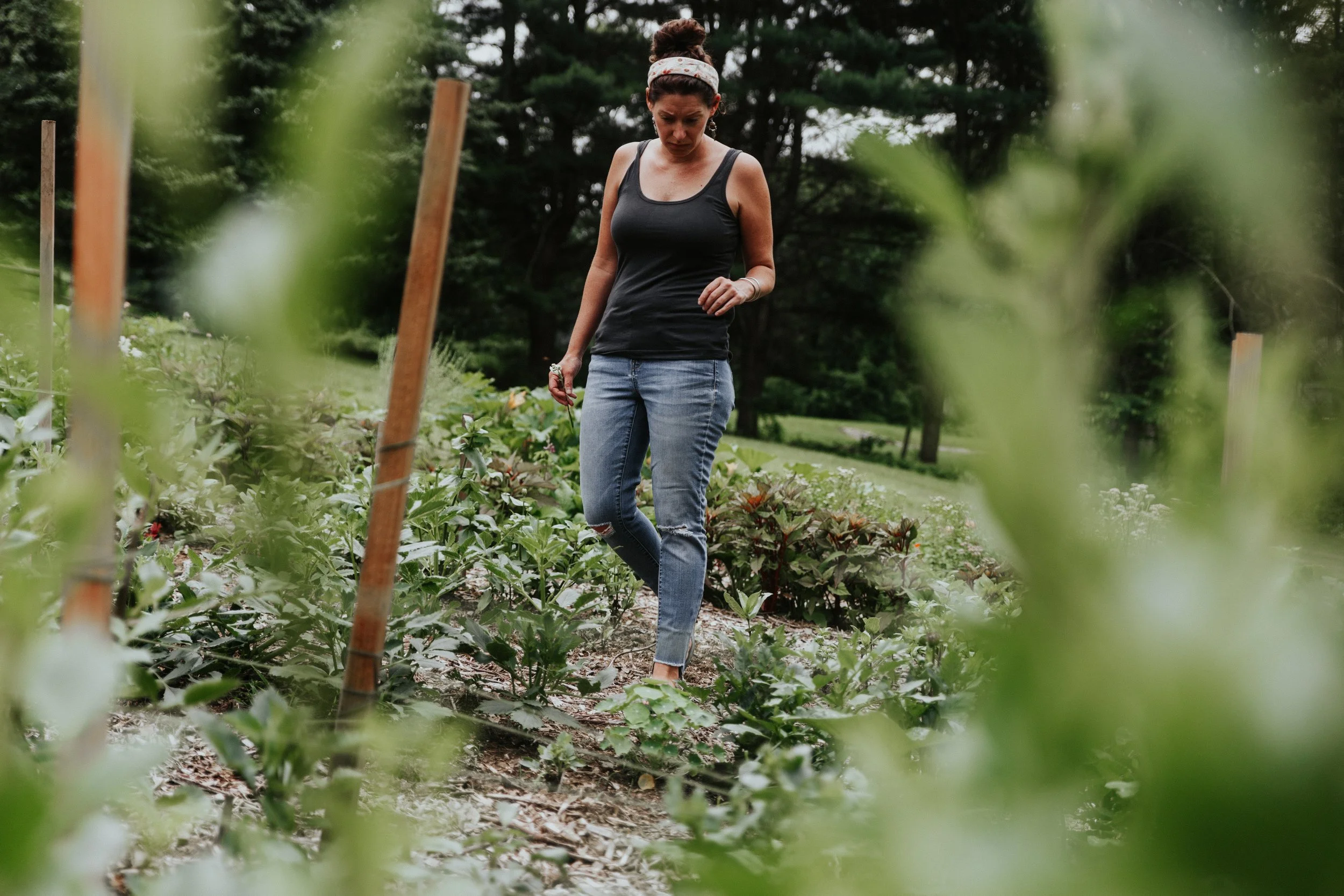 A woman with dark hair tied up, wearing a black tank top, blue jeans, and a white headband with red dots, is standing in a garden or farm with various leafy plants and supported by wooden stakes. She is looking down at the plants, with a serious or f
