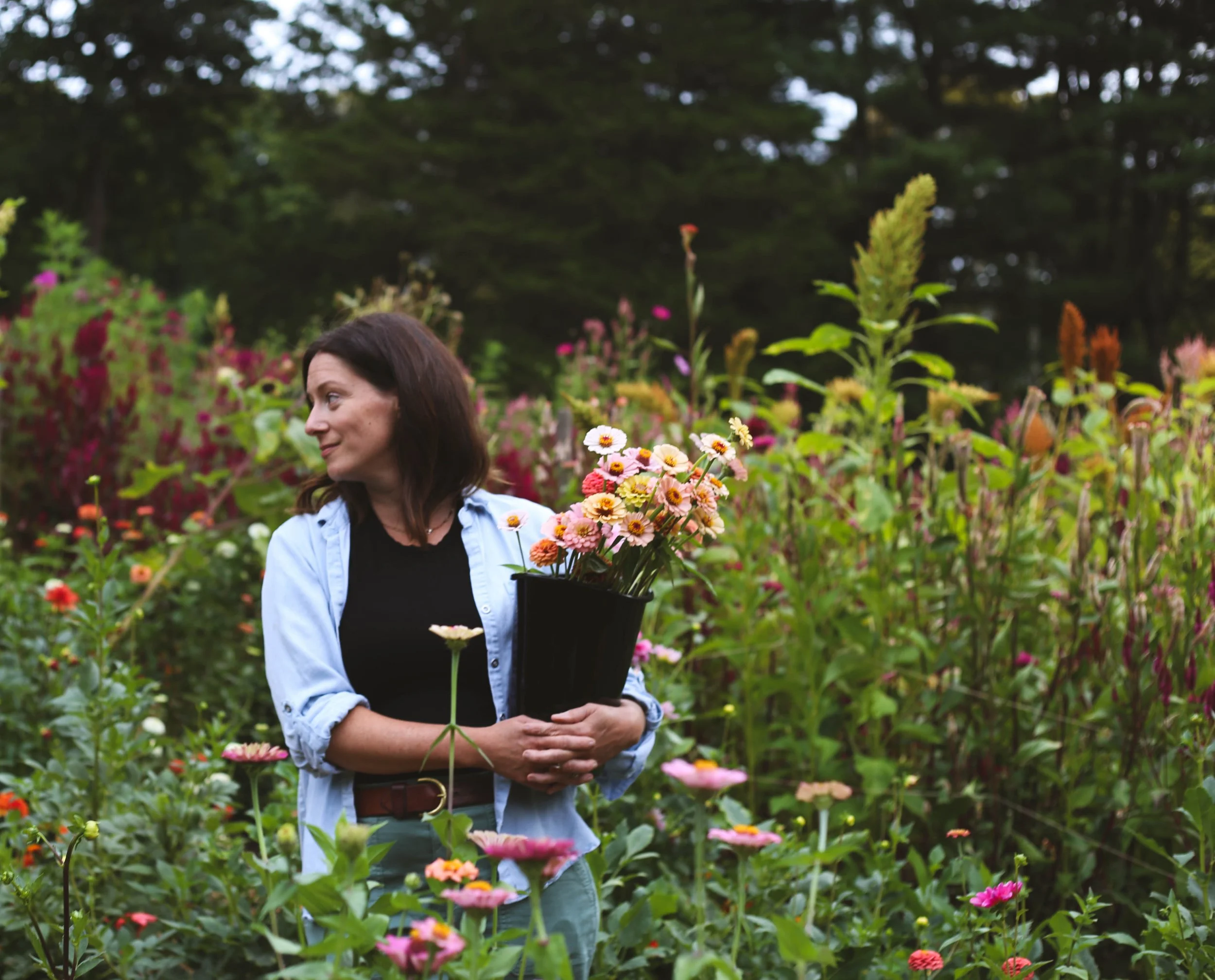 A woman with dark hair holding a black pot filled with pink, white, and yellow flowers in a garden with colorful blooming plants and dark trees in the background.