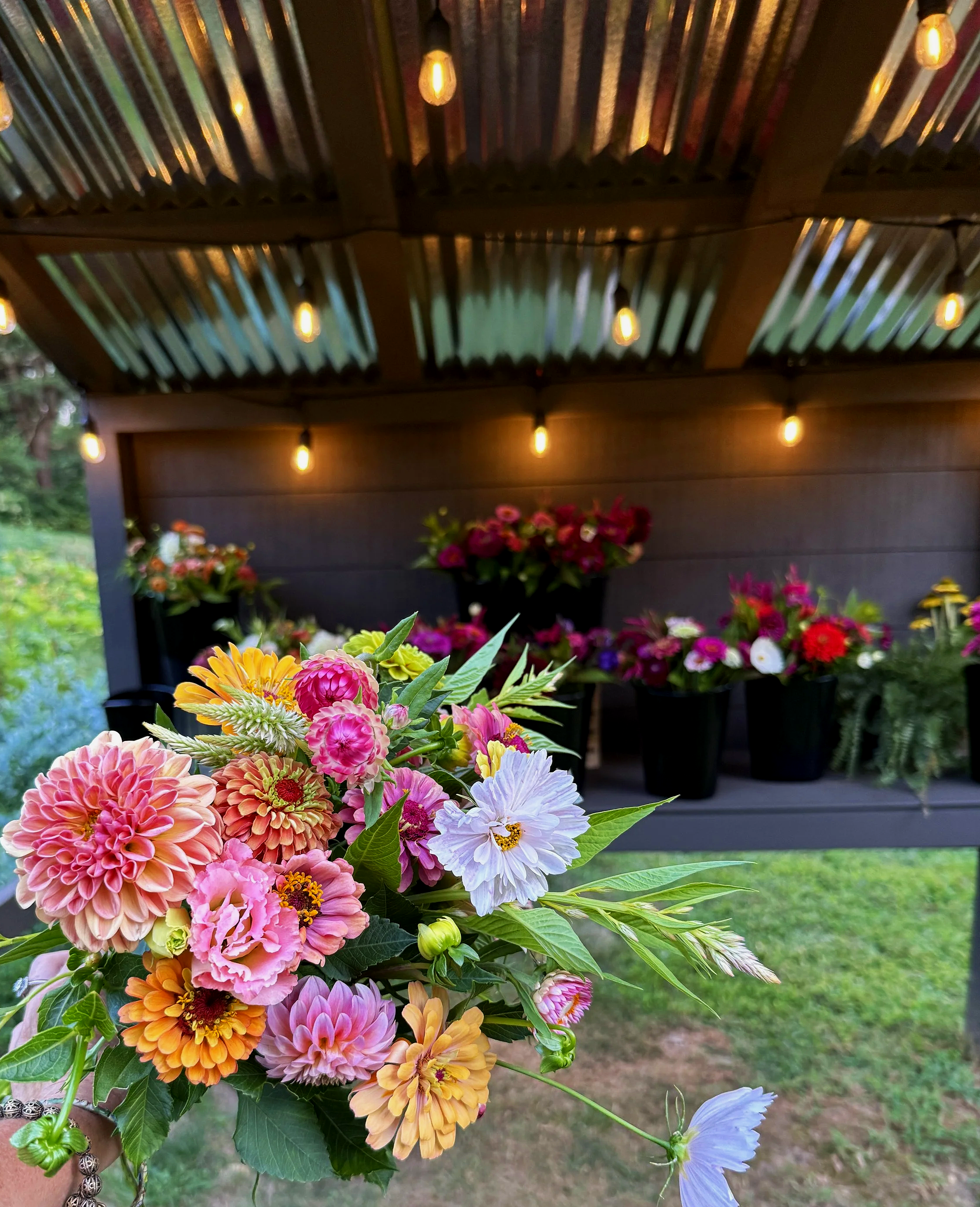 Colorful flower bouquet with pink, orange, yellow, and purple flowers, greenery, and a bun shape with a silver bracelet on the left, set against a background of a porch with string lights and a grassy area.