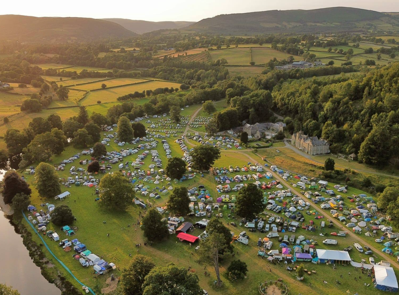 A scenic aerial view of a countryside festival with a large number of tents, parked cars, and trees set amidst green fields and rolling hills at sunset.