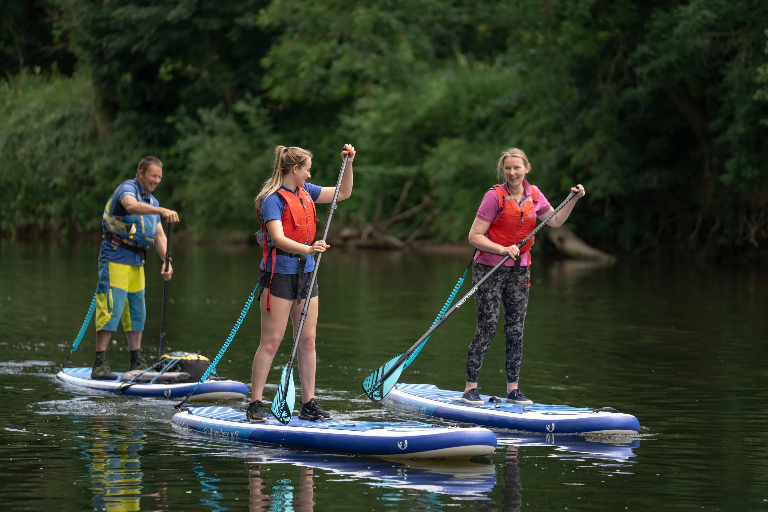Four people paddleboarding on a river surrounded by greenery, smiling and enjoying the activity.