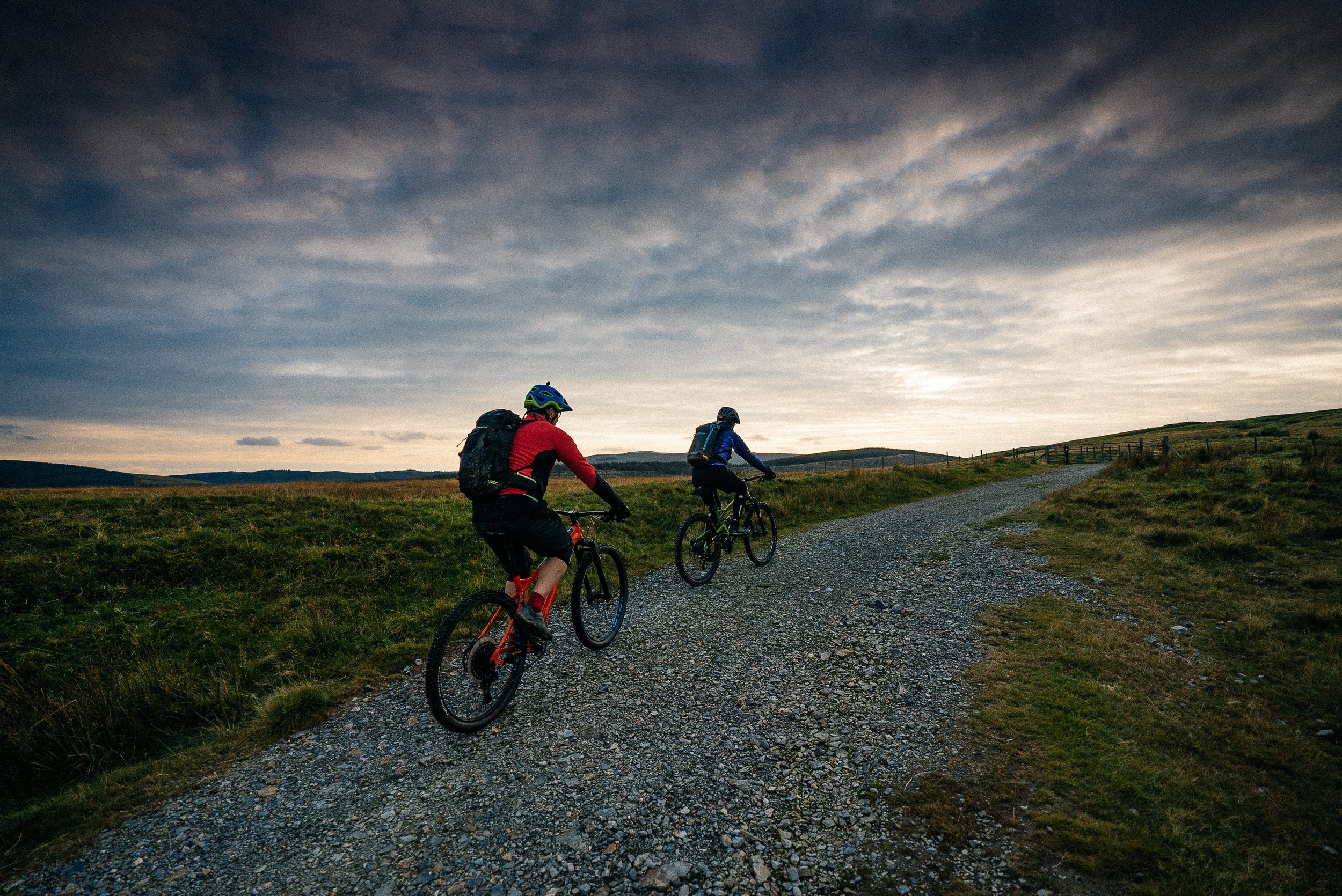 Two cyclists riding on a gravel path through a grassy landscape during sunset or dusk with a cloudy sky overhead.