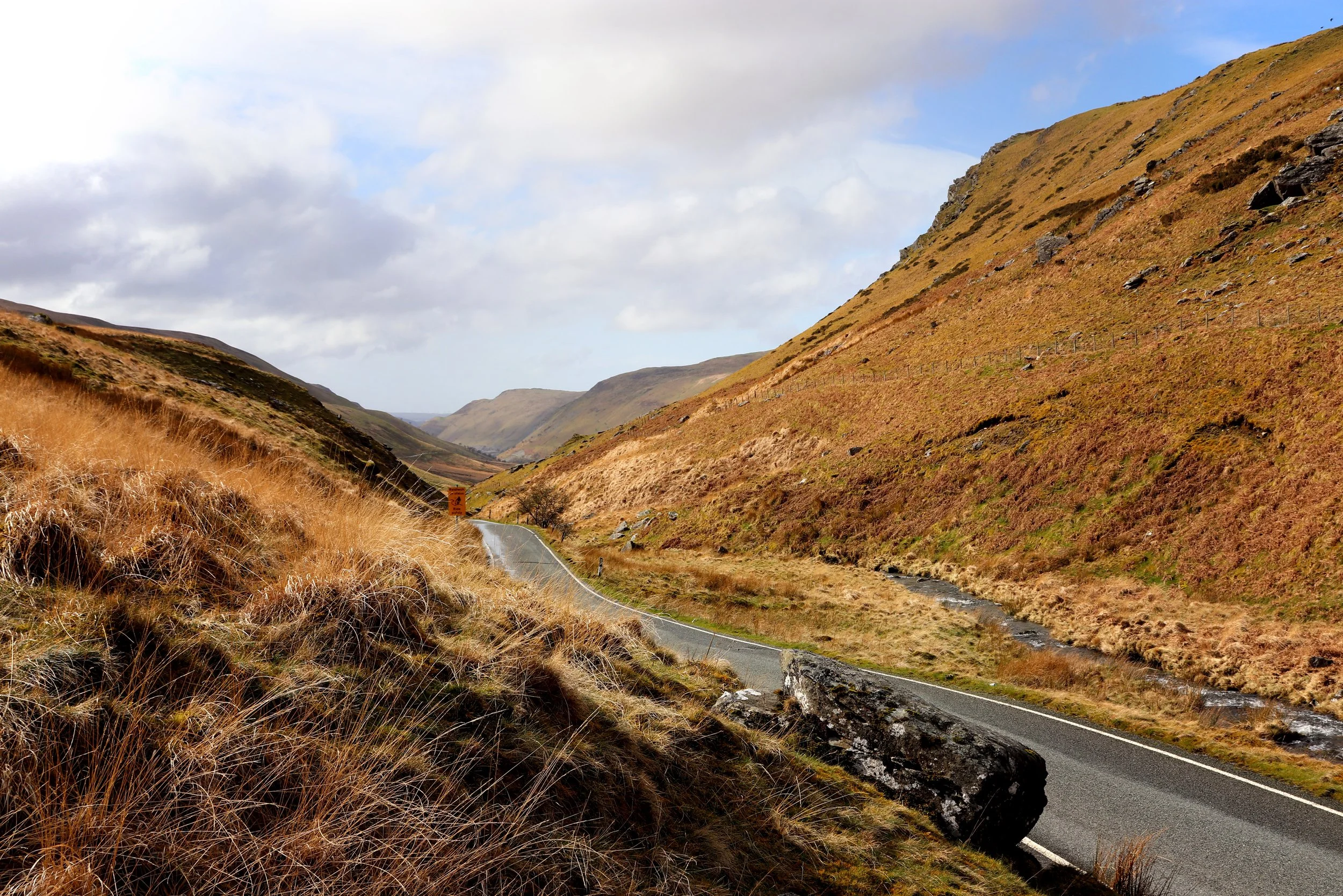 Scenic mountain road winding through grassy, rocky hills with cloudy sky overhead.