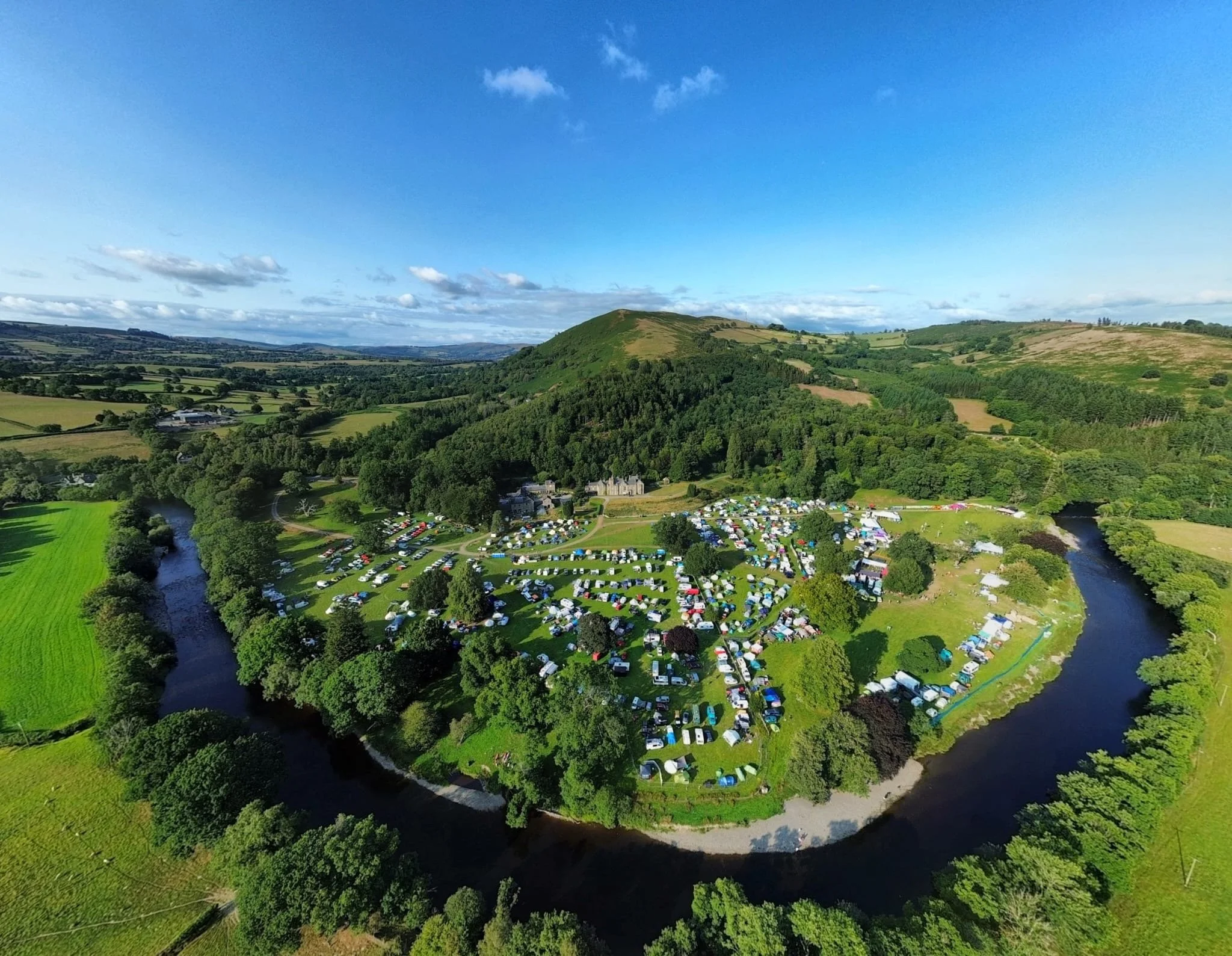 A scenic countryside landscape with rolling green hills, a river running through the valley, a large tent camping area, and a parking lot filled with cars. Bright blue sky with scattered clouds overhead.